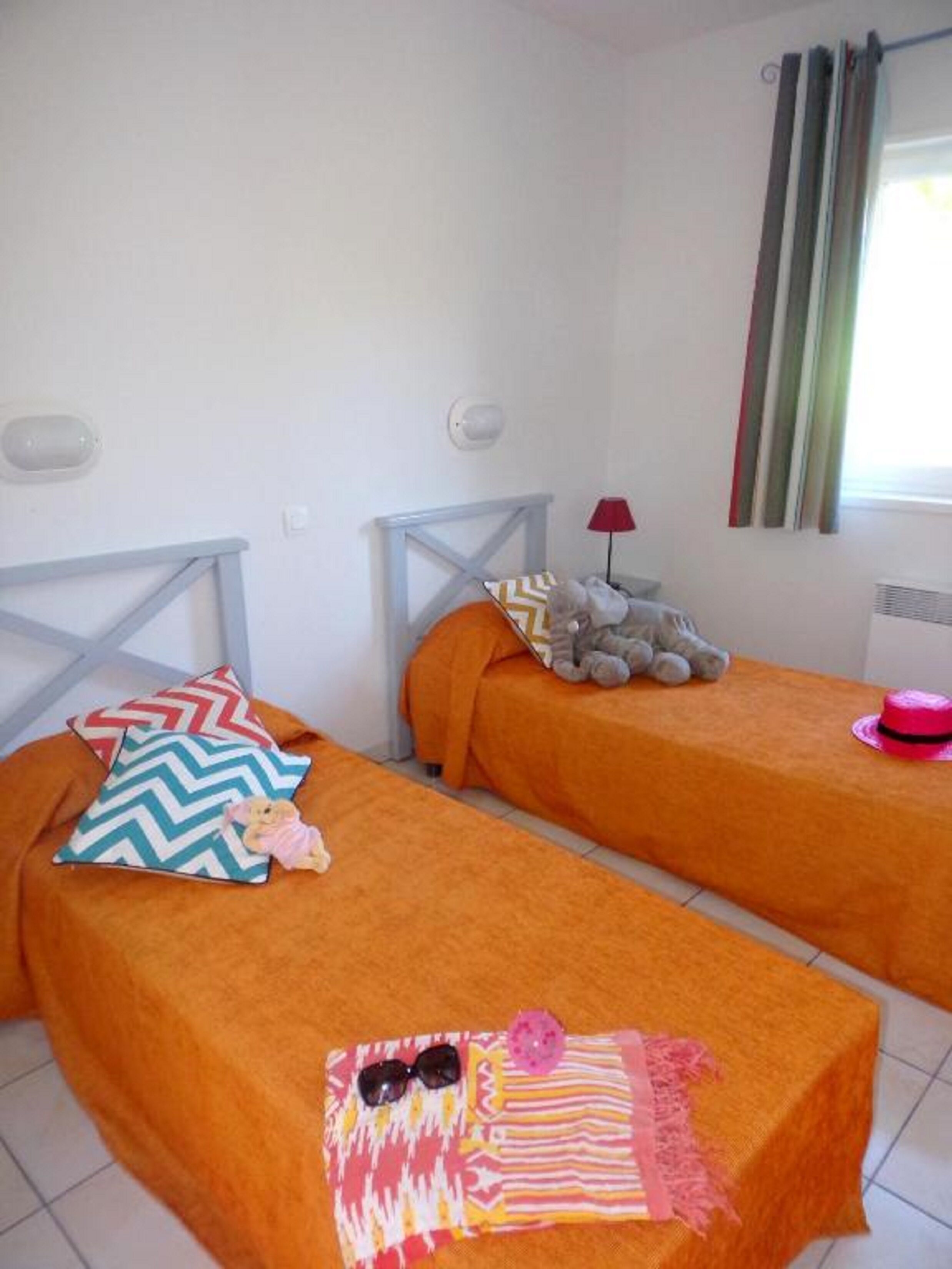 A room with two beds, each with an orange bedspread and a pillow. There is a window with curtains, a lamp, and a stuffed toy on one bed.