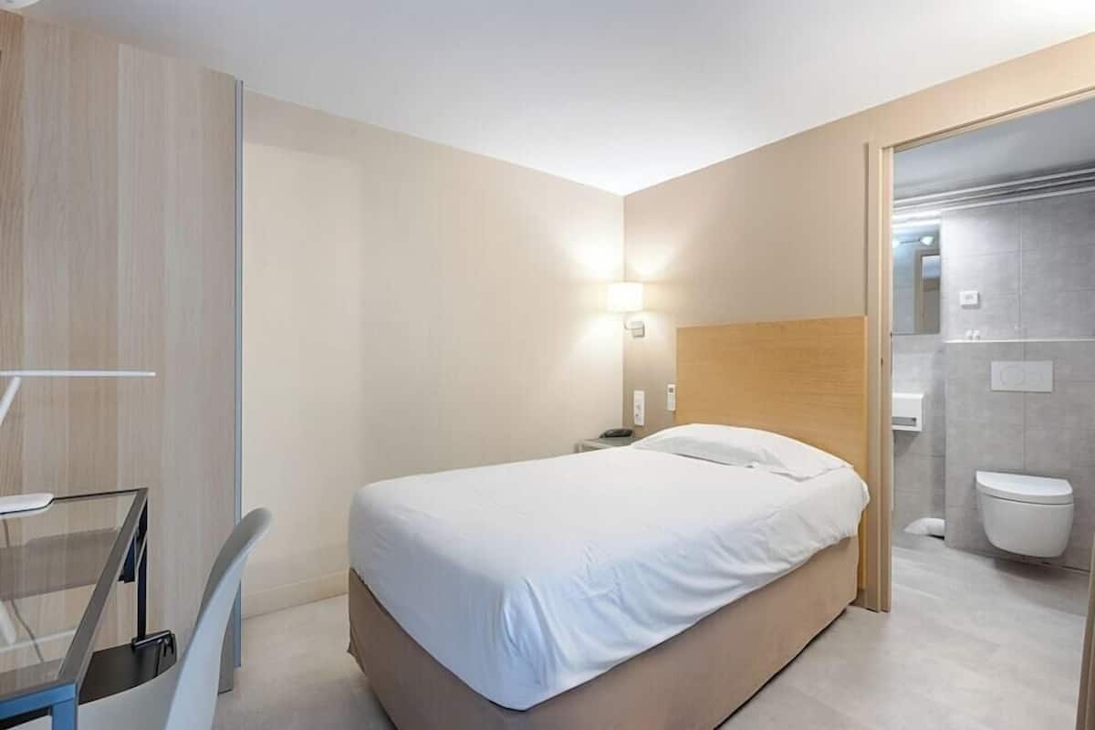 A modern hotel room with a bed, bedside table, and a glass desk with a chair.