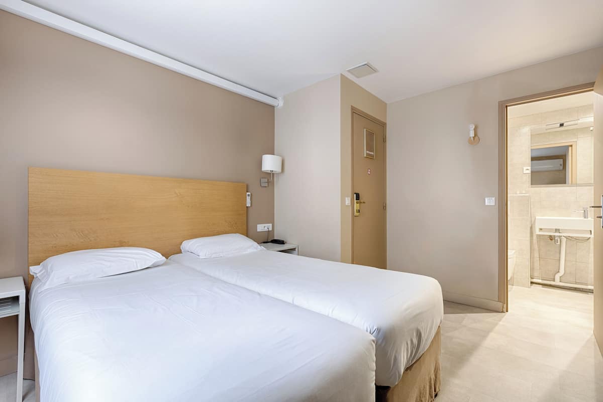 A hotel room with two beds, a nightstand, a lamp, and a bathroom visible through an open door.