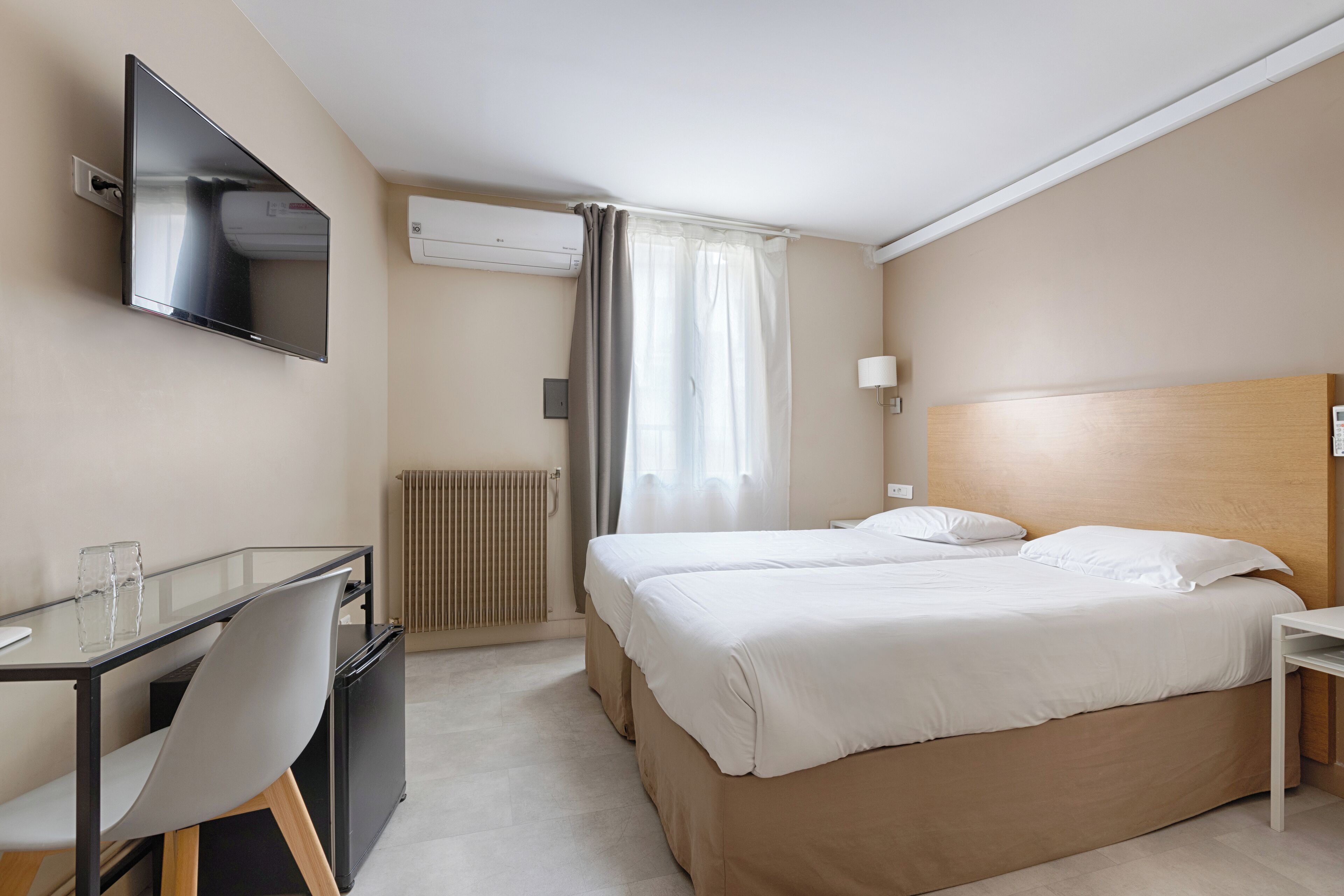 A hotel room with a bed, a desk with a chair, a TV mounted on the wall, and an air conditioning unit.