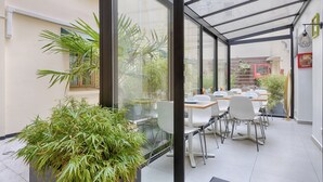 Een moderne, overdekte eetruimte met glazen wanden, witte stoelen en een grote potplant.