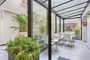 A modern indoor dining area with glass walls, white chairs, and a large potted plant.