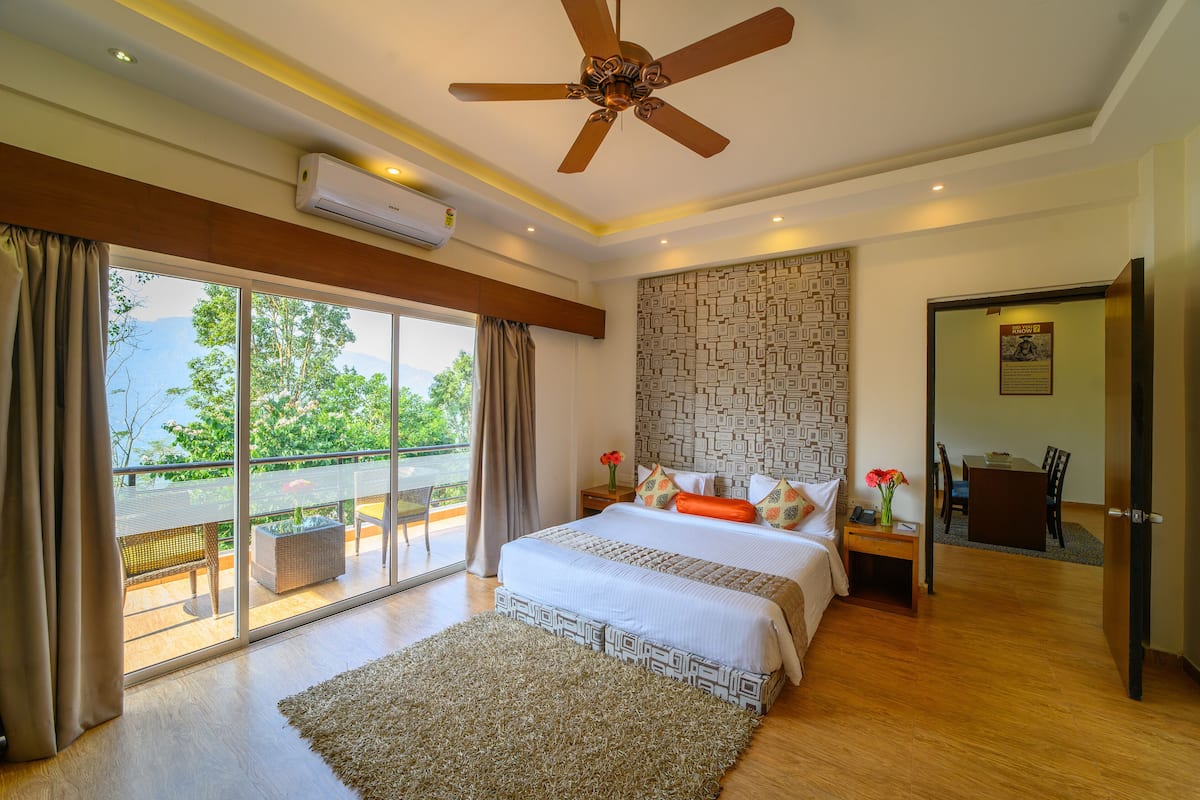Une chambre avec un grand lit, un ventilateur de plafond, un appareil de climatisation, un mur décoré d’un motif, une vue sur l’extérieur par une porte-fenêtre coulissante et une petite table sur laquelle est posé un vase de fleurs.