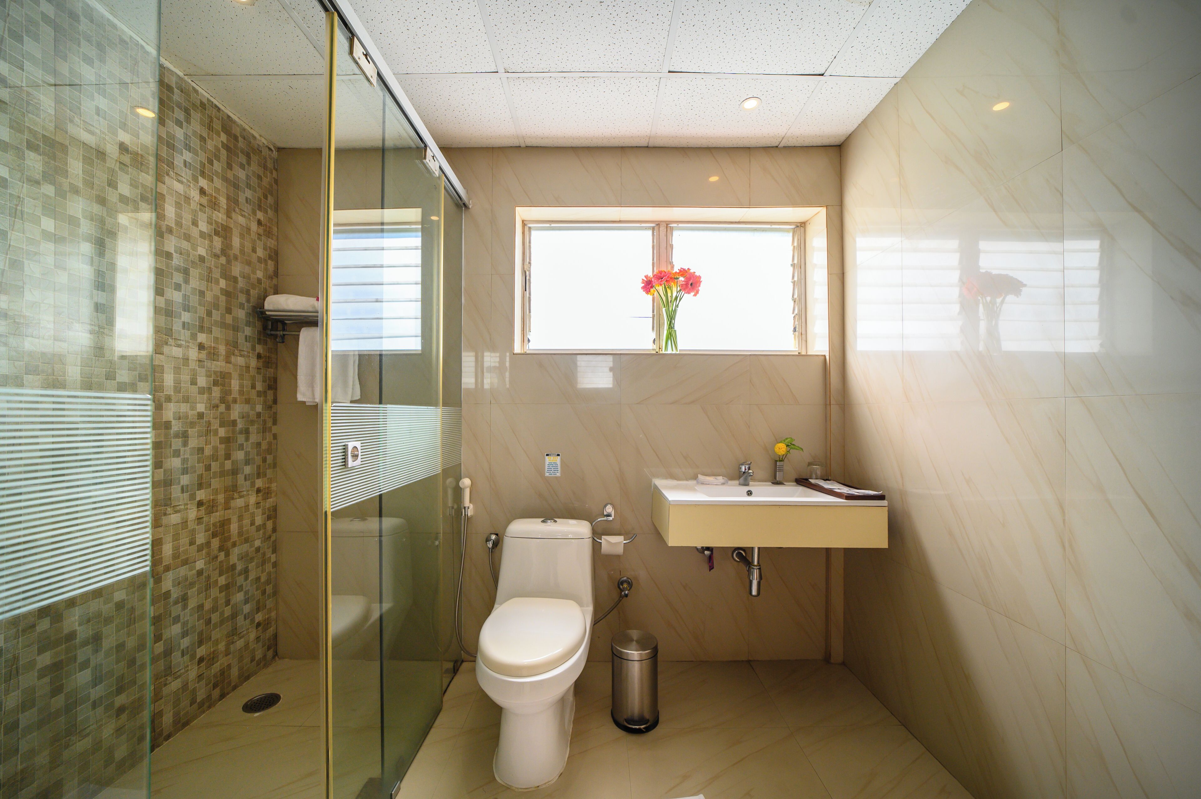 Un baño moderno con cabina de ducha de vidrio, inodoro, lavamanos y ventana con un florero.
