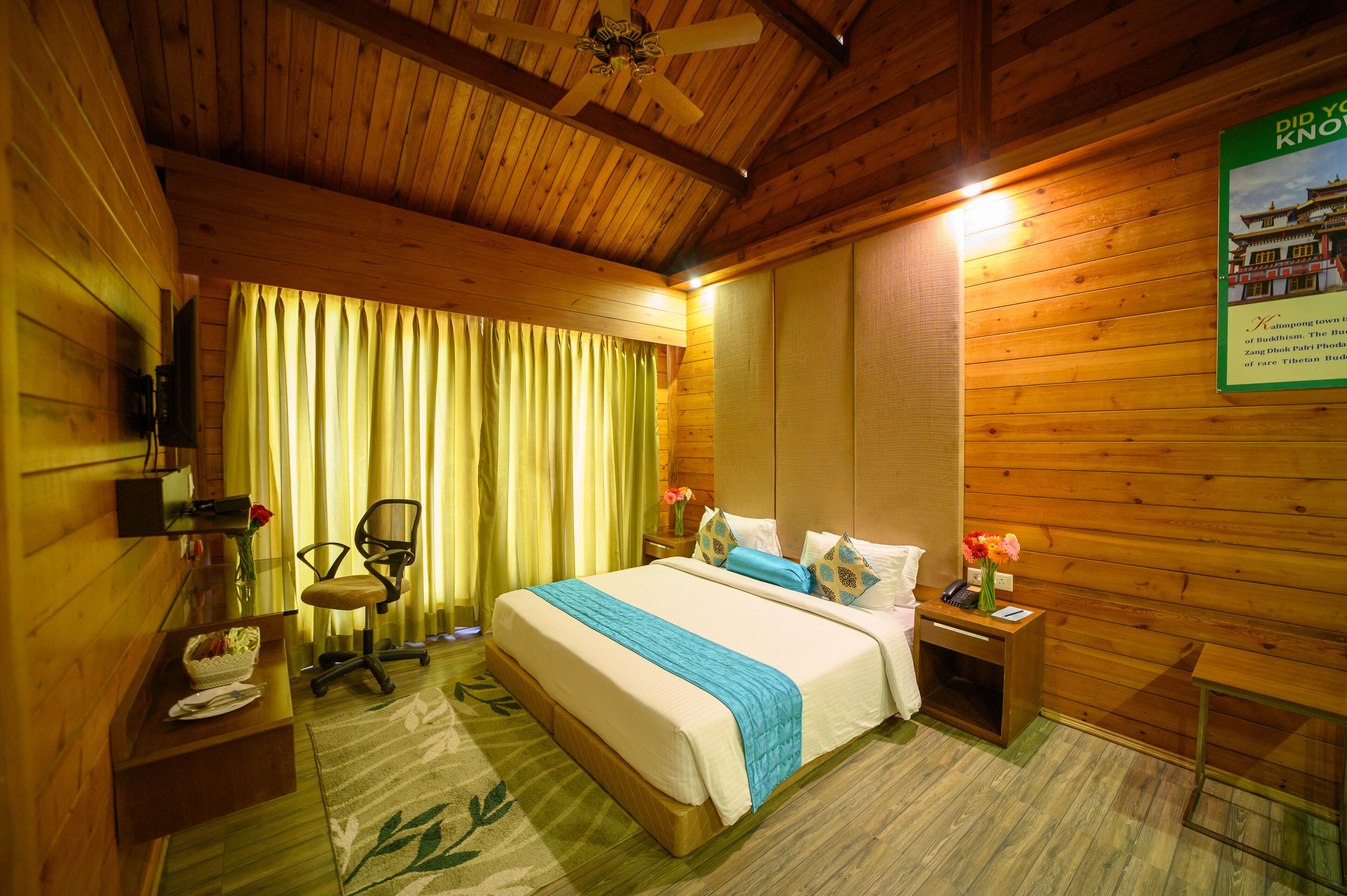 A wooden-themed hotel room with a bed, a desk, a chair, a nightstand, and a wall-mounted TV.