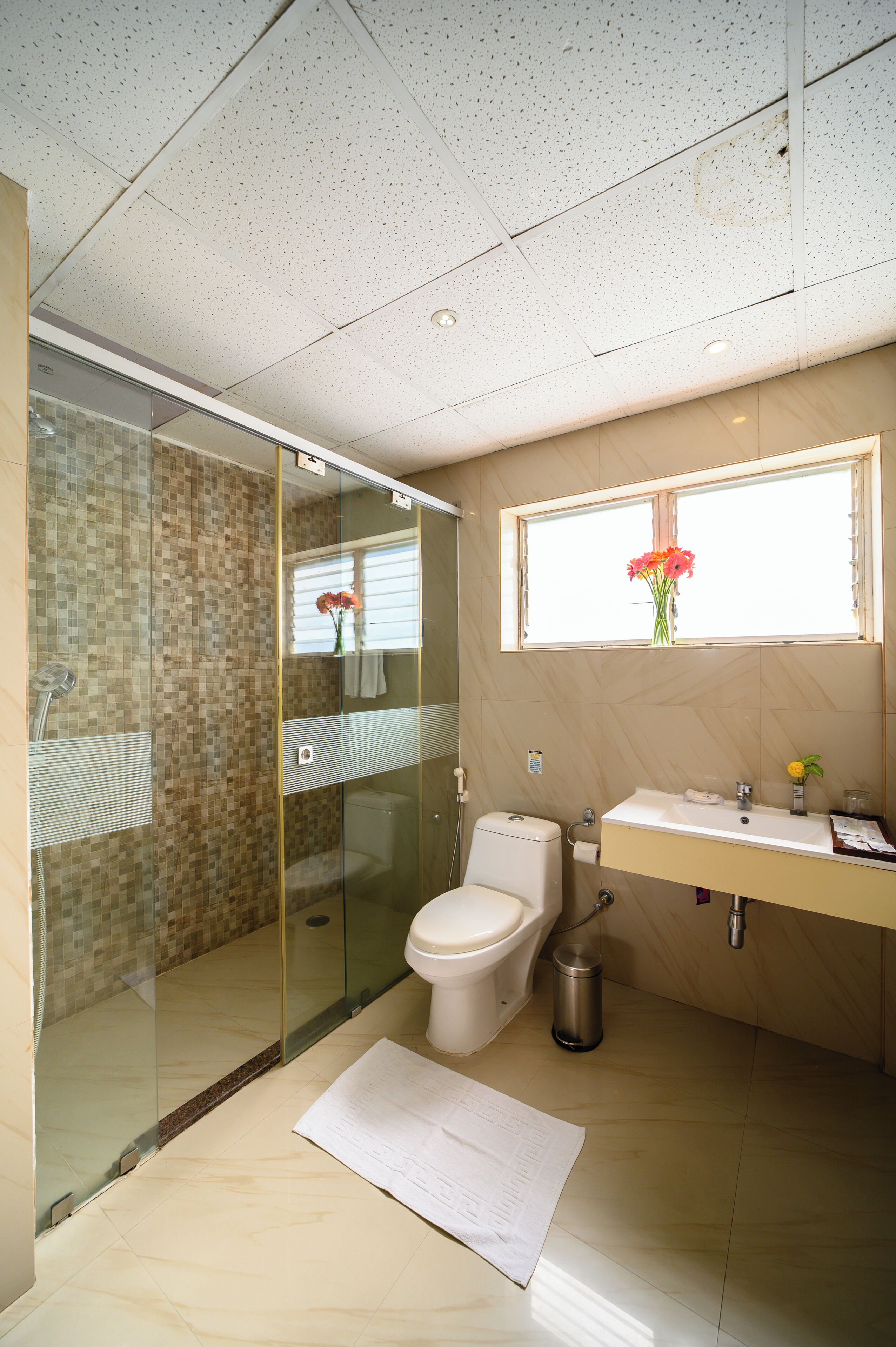 A modern bathroom with a glass shower enclosure, a toilet, a sink with a mirror, and a window with a flower vase.