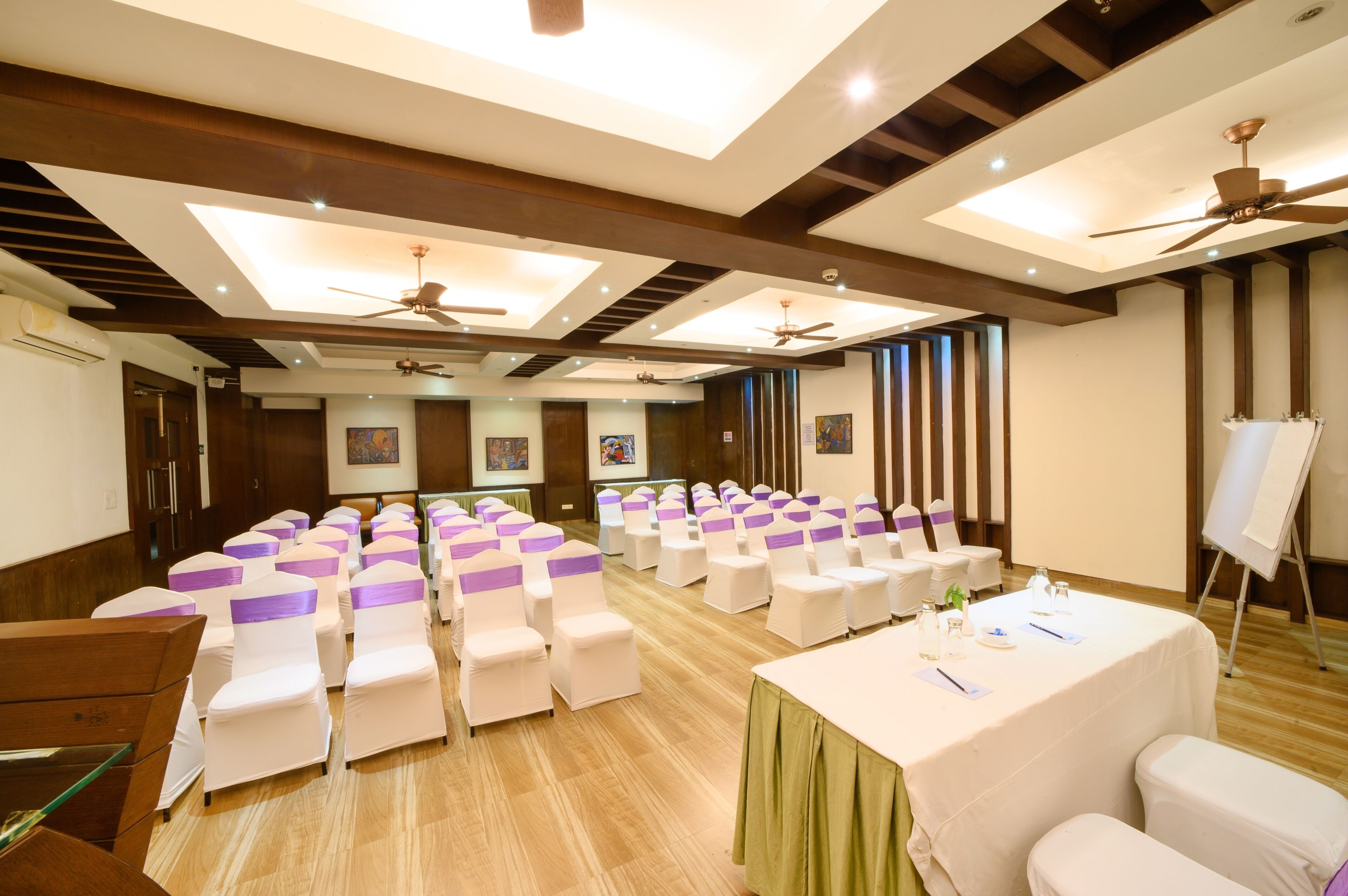 Une salle de conférence avec des rangées de chaises blanches, une table recouverte d’une nappe verte et des ventilateurs de plafond.