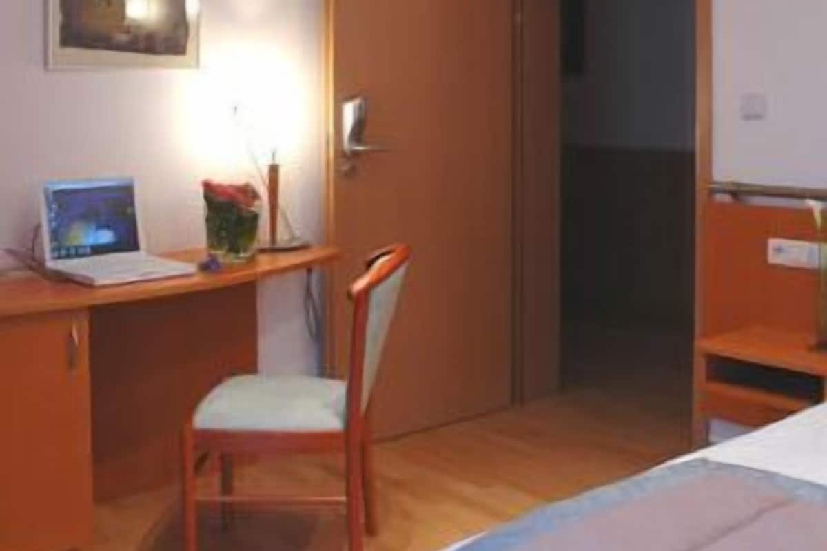 A bedroom with a bed, a desk with a laptop, a chair, and a door.