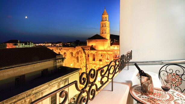 A balcony with a view of a historic building with a tower, a metal railing, a table with a bottle and glass, and a night sky with a moon. - La Porta Luxury Rooms (Split)
