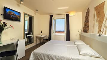 A hotel room with a bed, a desk, a TV, a window with a view, and a painting on the wall.