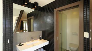 Een moderne badkamer met een wit wastafelmeubel, donkere betegelde wanden, een grote spiegel en een douchecabine met glazen wanden.