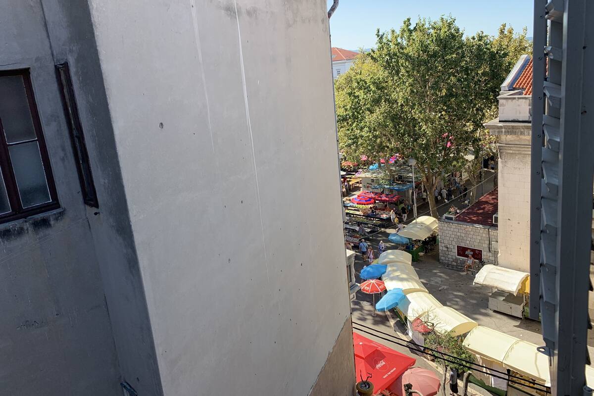 Vista de un mercado callejero con puestos diversos y sombrillas, rodeado de edificios y árboles.