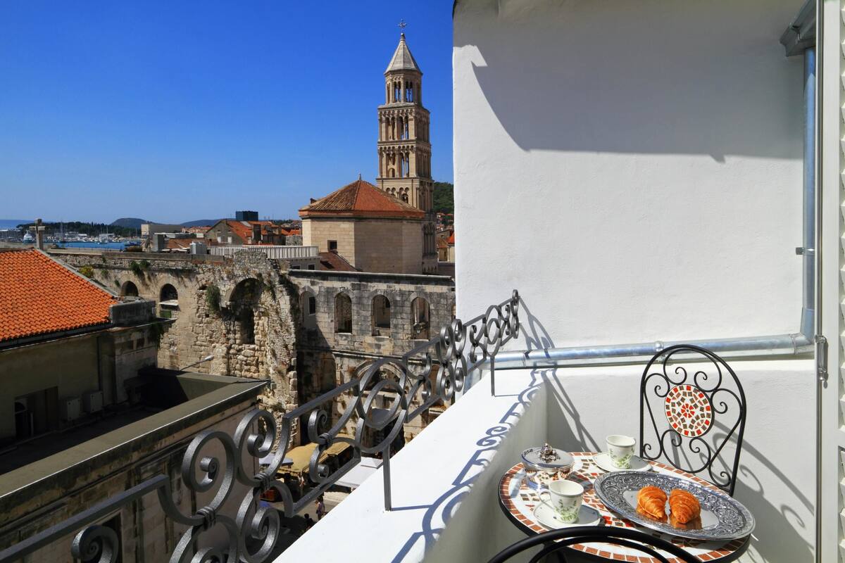 Un balcón con una mesa dispuesta para dos personas, con vista a un pueblo histórico y una torre destacada.
