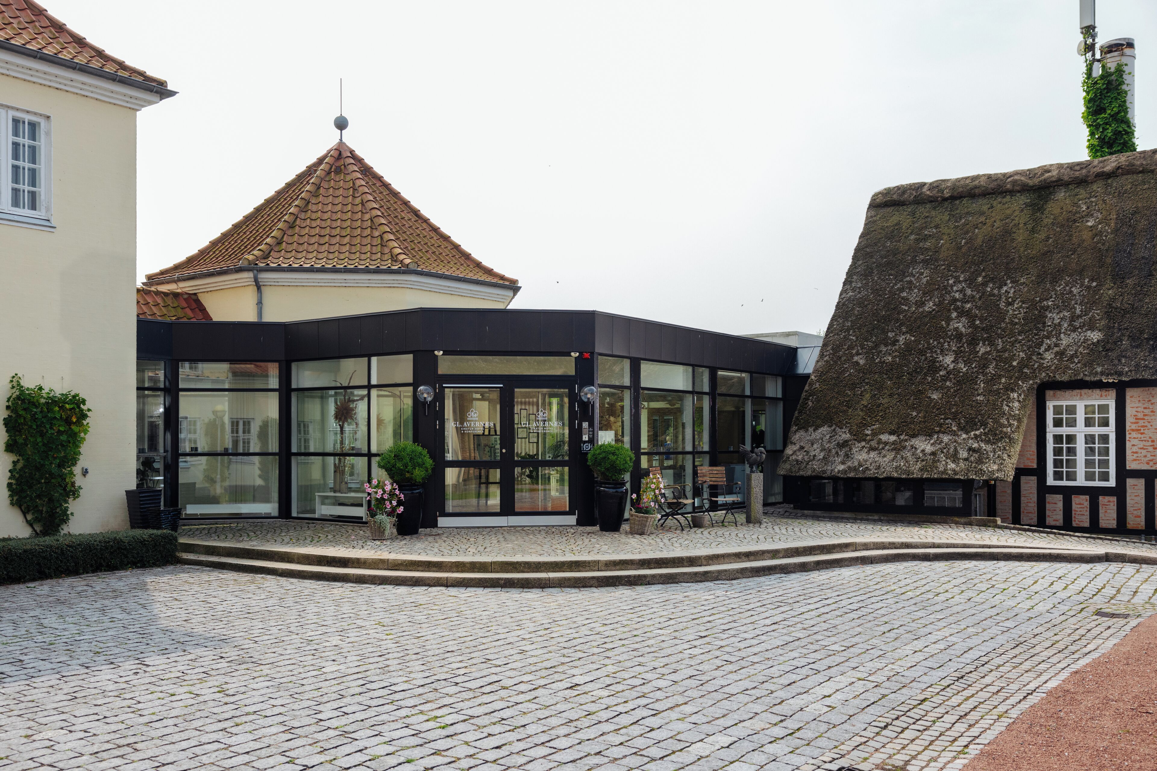 A building with a modern glass entrance, a traditional thatched roof structure, and cobblestone paving.