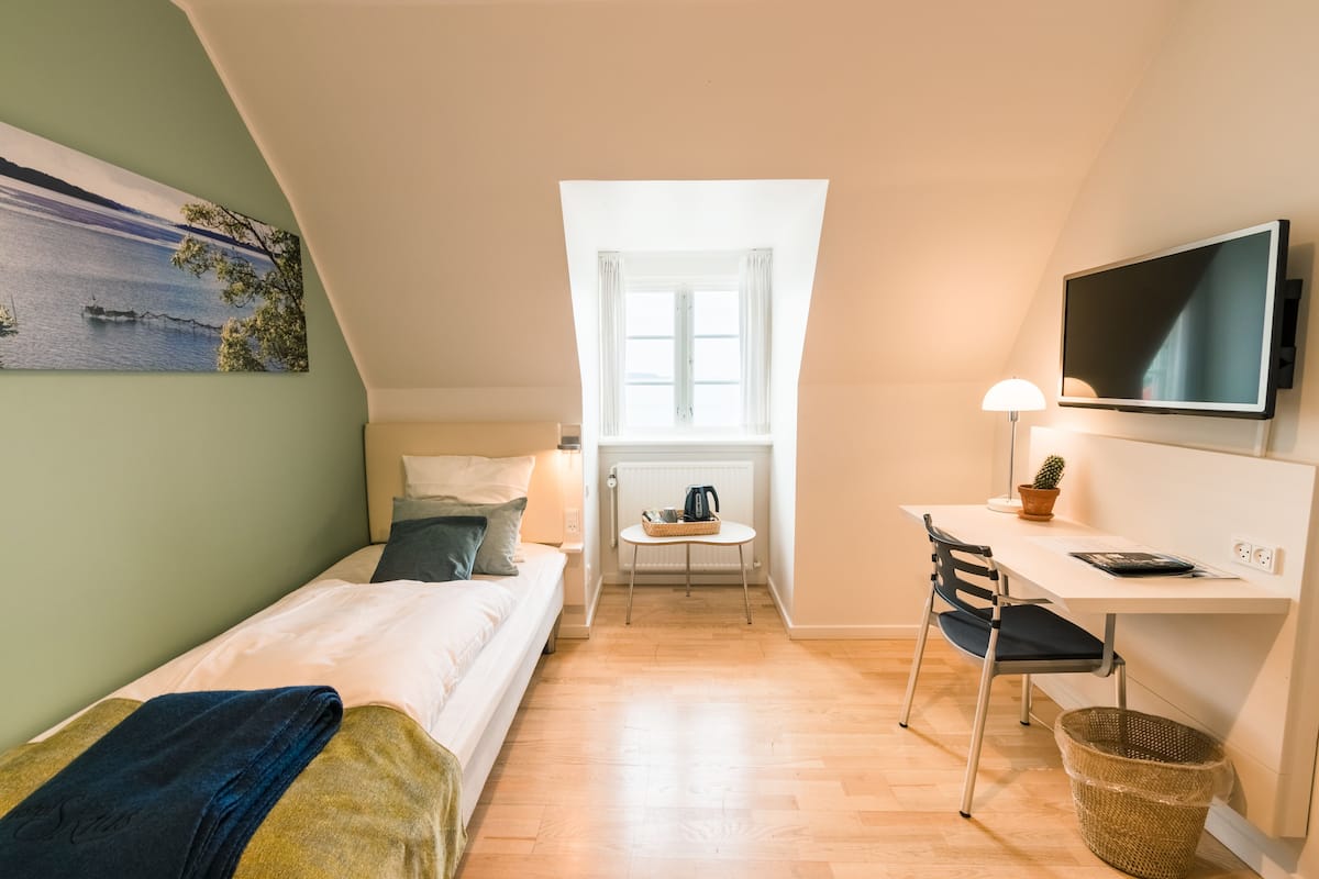 Una habitación pequeña y luminosa con cama, escritorio, silla, televisor y ventana con cortinas.