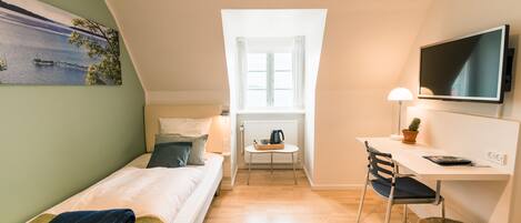 Una habitación pequeña y luminosa con cama, escritorio, silla, televisor y ventana con cortinas.