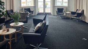 Een moderne hotelentree met een zwarte leren bank, twee fauteuils, een klein rond tafeltje en een potplant. Grote ramen bieden uitzicht op de zee.