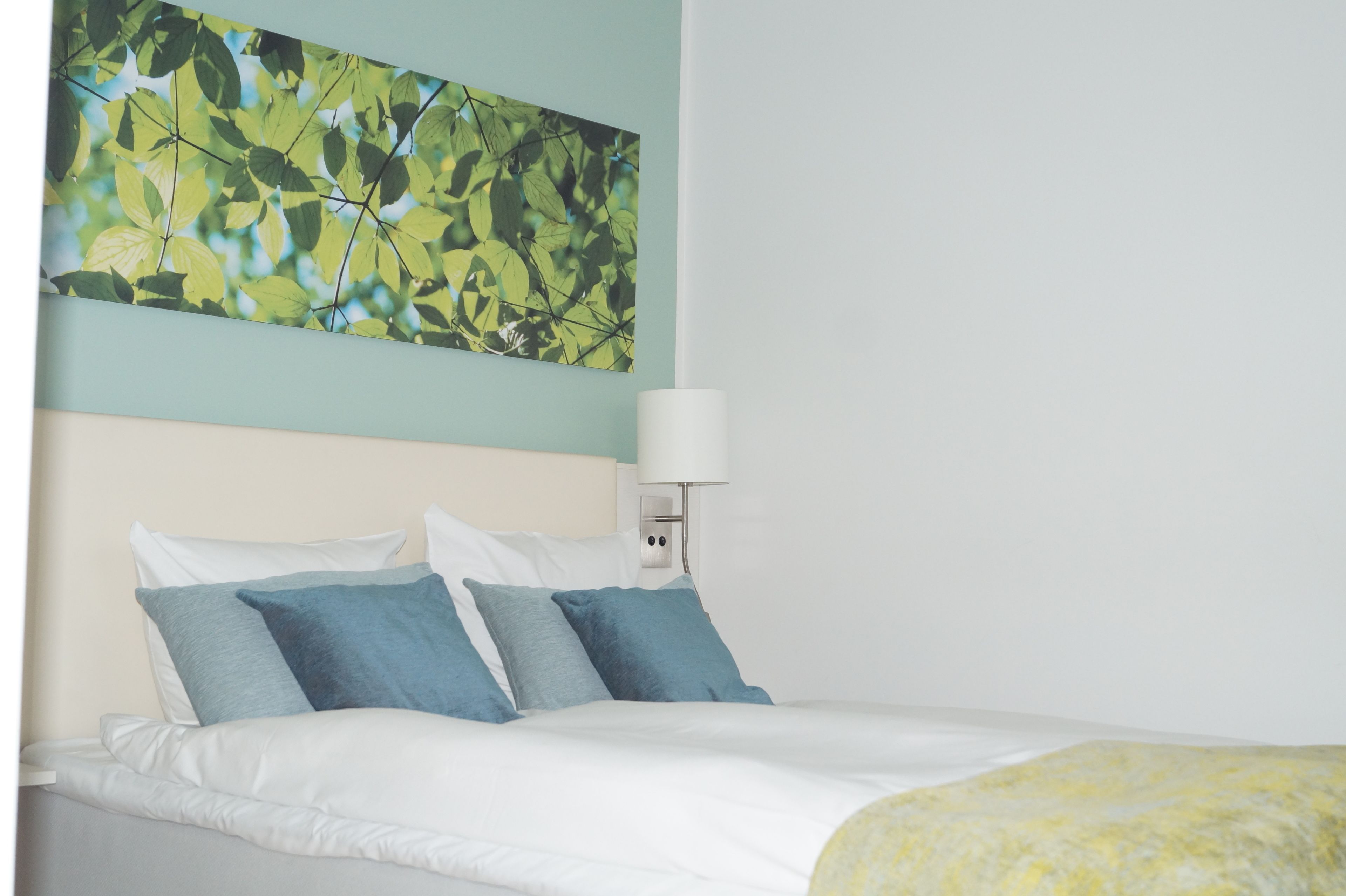 Una cama bien hecha con ropa de cama blanca y azul, una lámpara fijada a la pared y un cuadro abstracto grande sobre la cama.