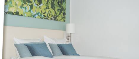 A neatly made bed with white and blue bedding, a wall-mounted lamp, and a large abstract painting above the bed.