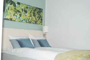 Una cama bien hecha con ropa de cama blanca y azul, una lámpara fijada a la pared y un cuadro abstracto grande sobre la cama.