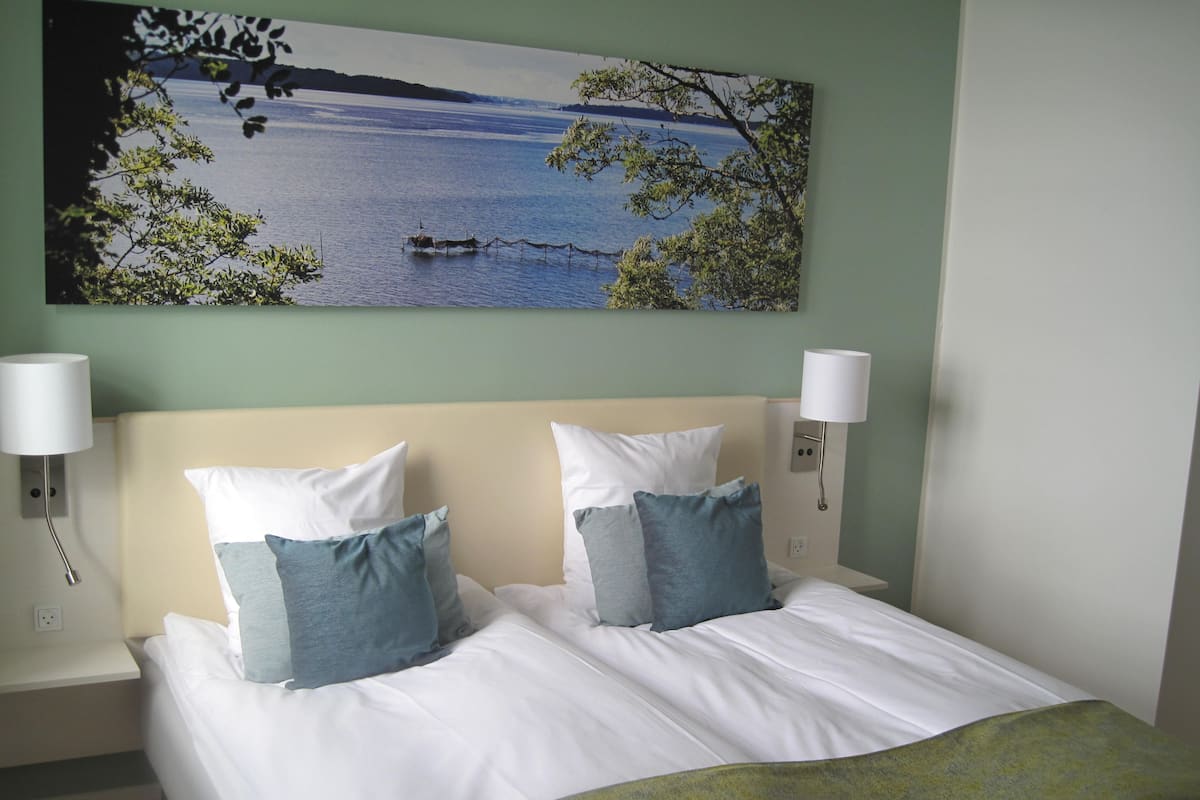 Una cama bien hecha con ropa de cama blanca y azul, dos lámparas de pared y un cuadro con un paisaje de lago sobre la cama.