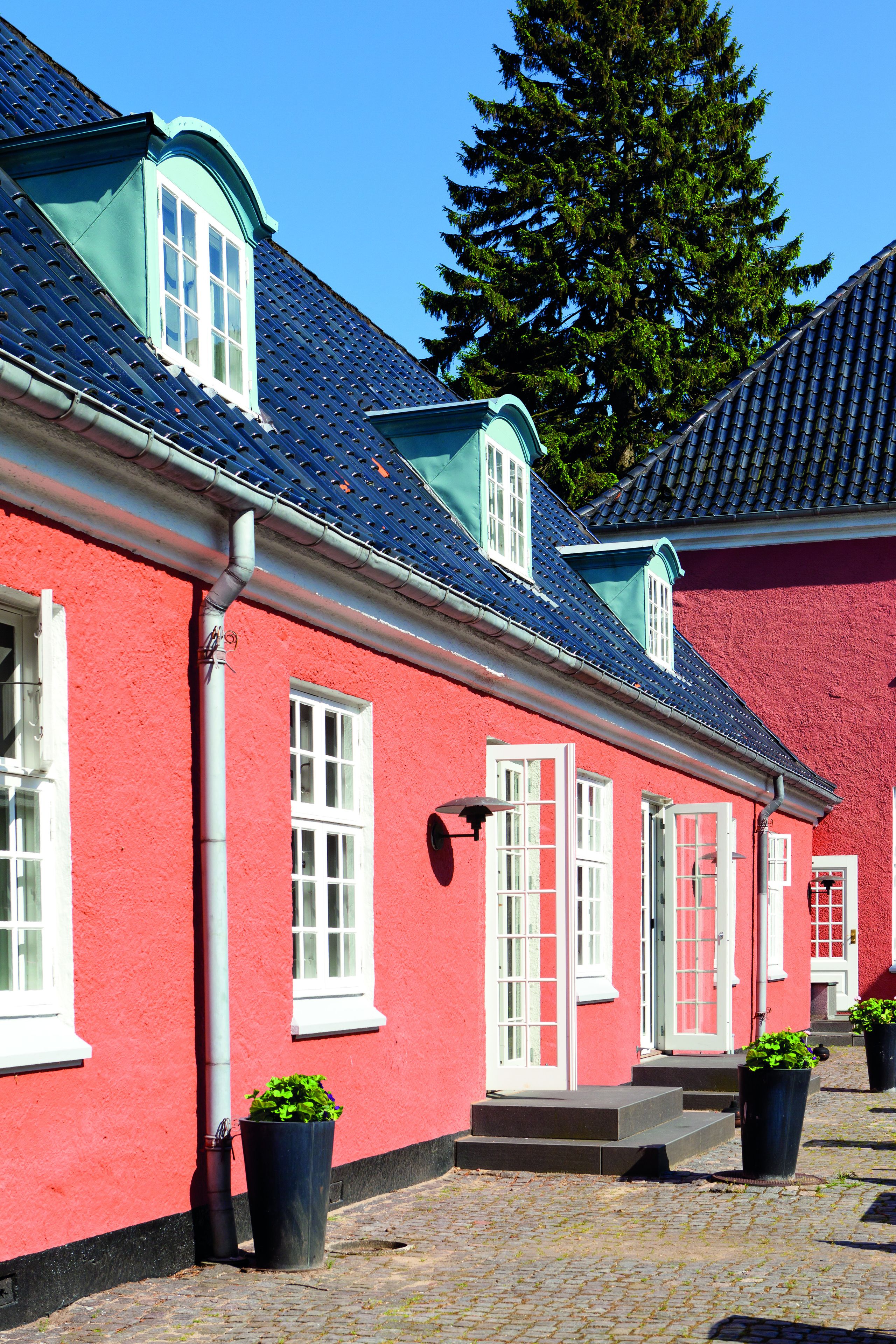 Um edifício vermelho com janelas e portas brancas, telhado em telhas e plantas em vasos no chão.