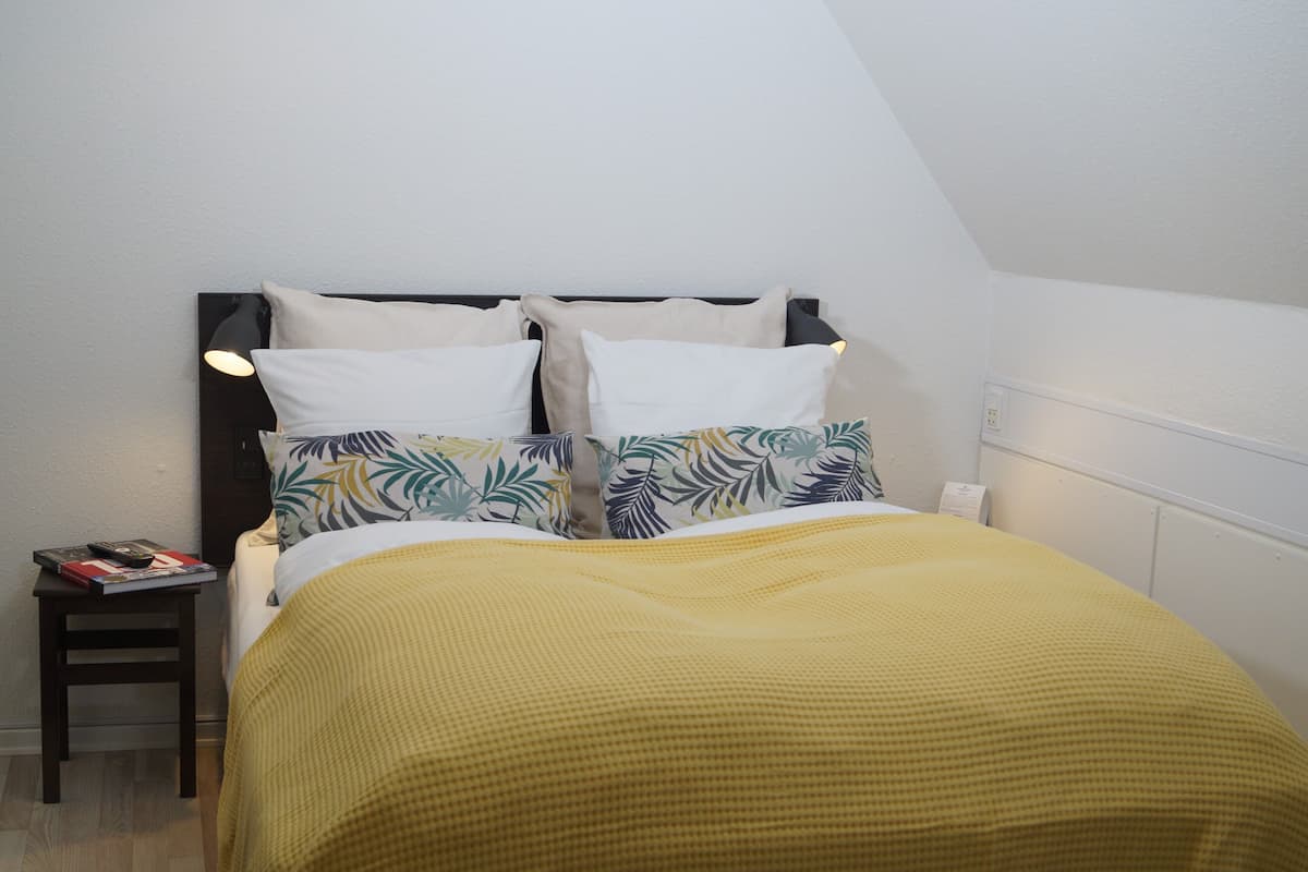 Ein ordentlich gemachtes Bett mit einer gelben Decke, ein Nachttisch mit einem Buch und einer Fernbedienung sowie eine Wandlampe.