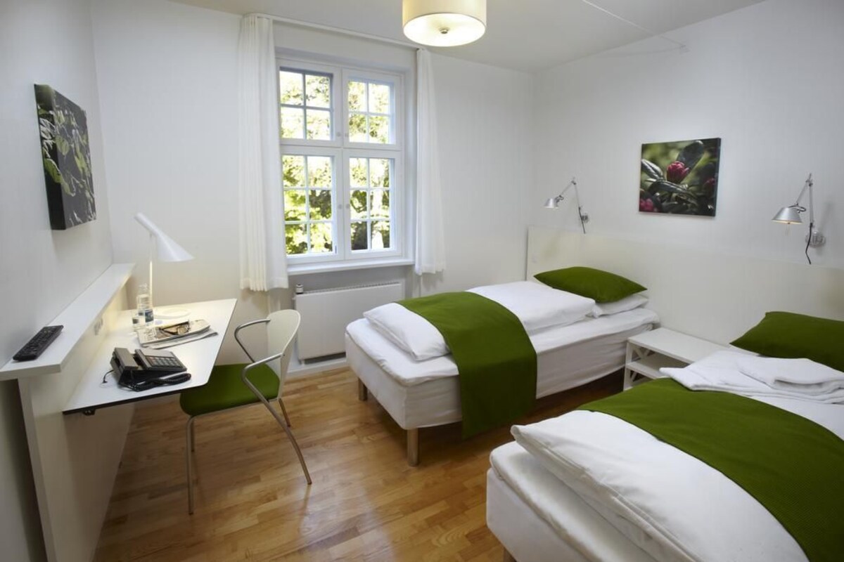 A hotel room with a desk, two beds, a chair, a window with curtains, and artwork on the walls.