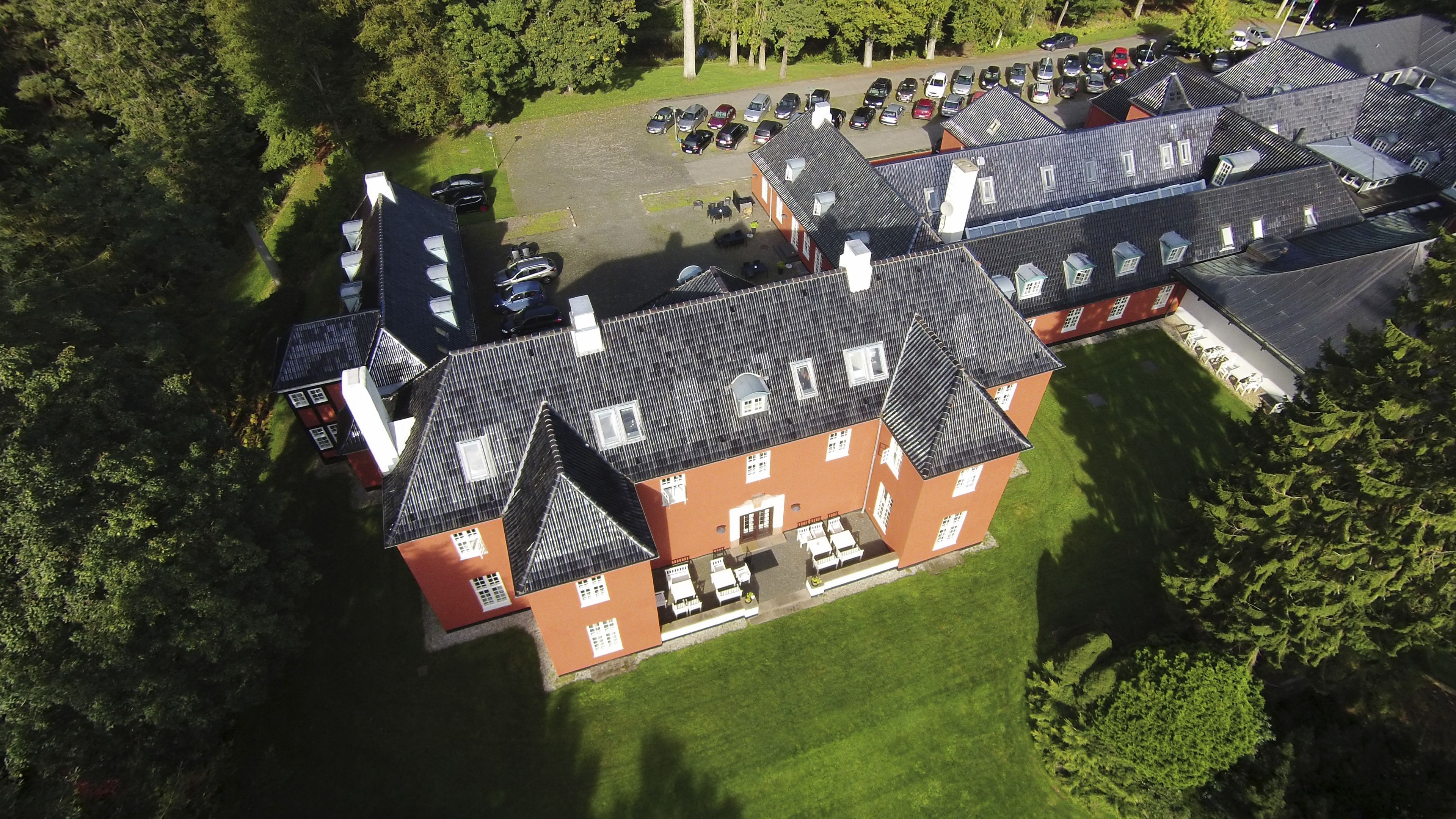 An aerial view of a large, multi-story building with a red facade and dark roof. There is a parking lot with several cars and a small outdoor seating area.