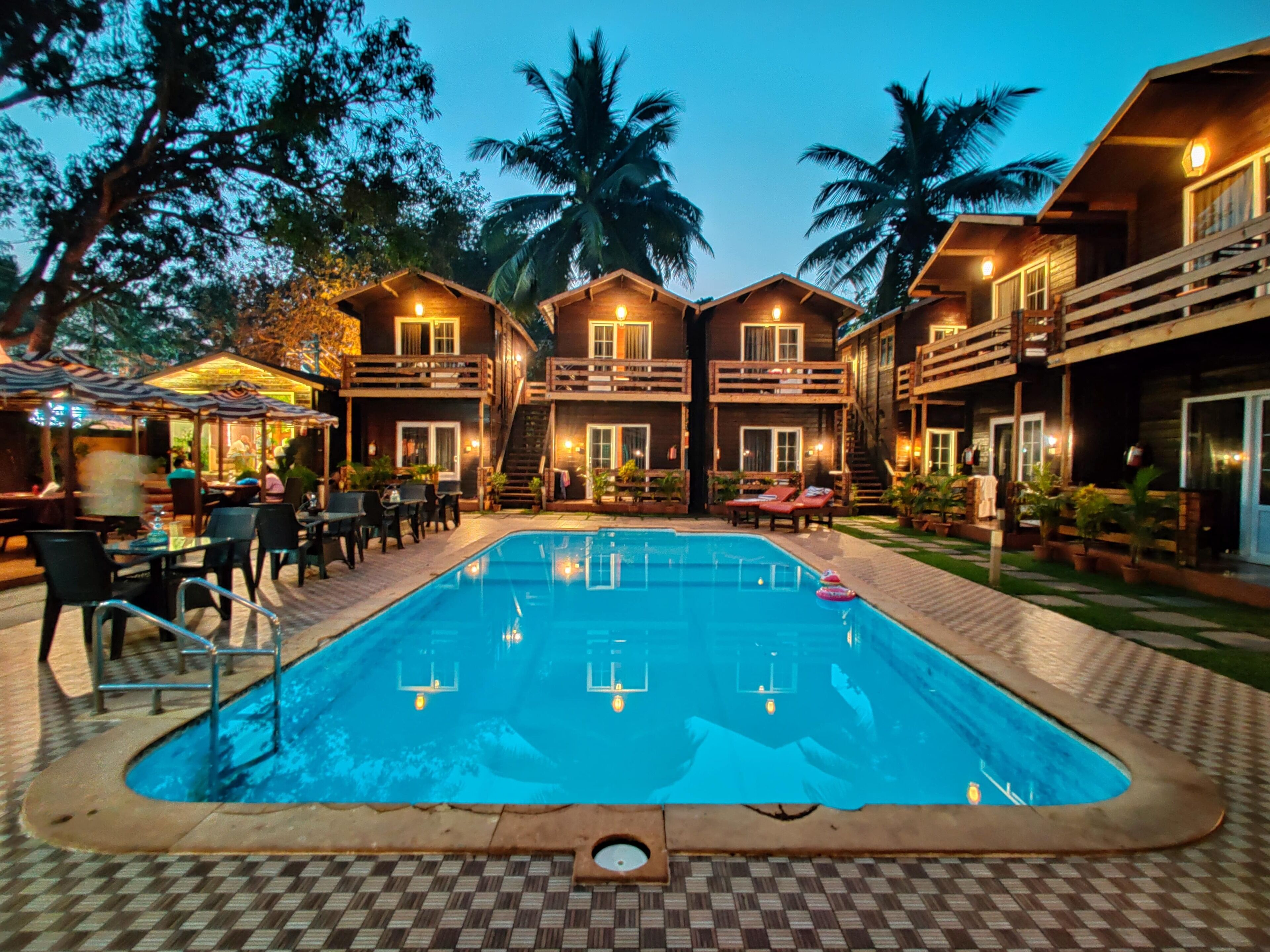 A pool area with a checkered floor, surrounded by wooden buildings with balconies, and palm trees in the background. - La Vie Woods (Calangute)