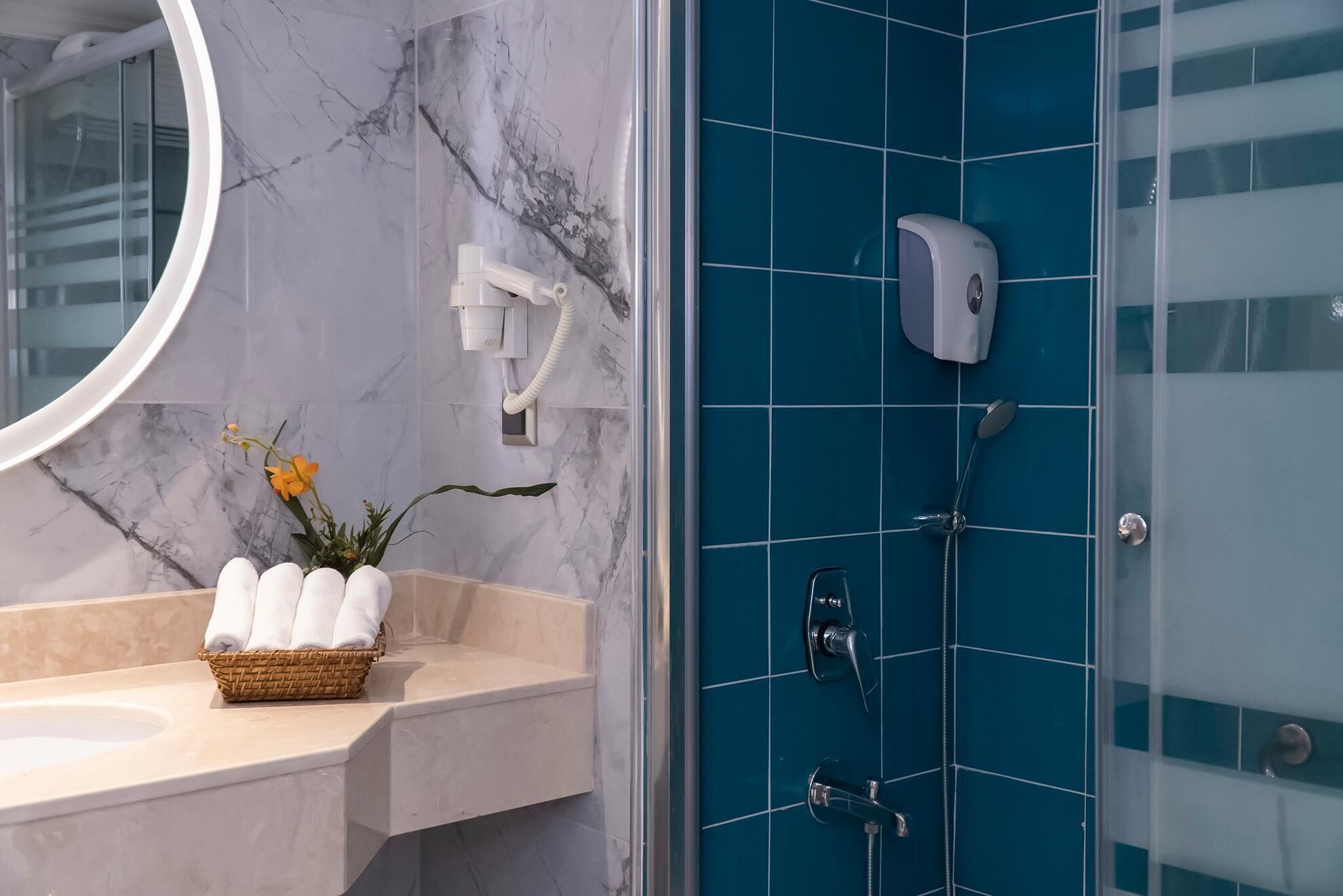 A bathroom with a marble vanity, a basket of towels, a hairdryer, and a shower with blue tiles.