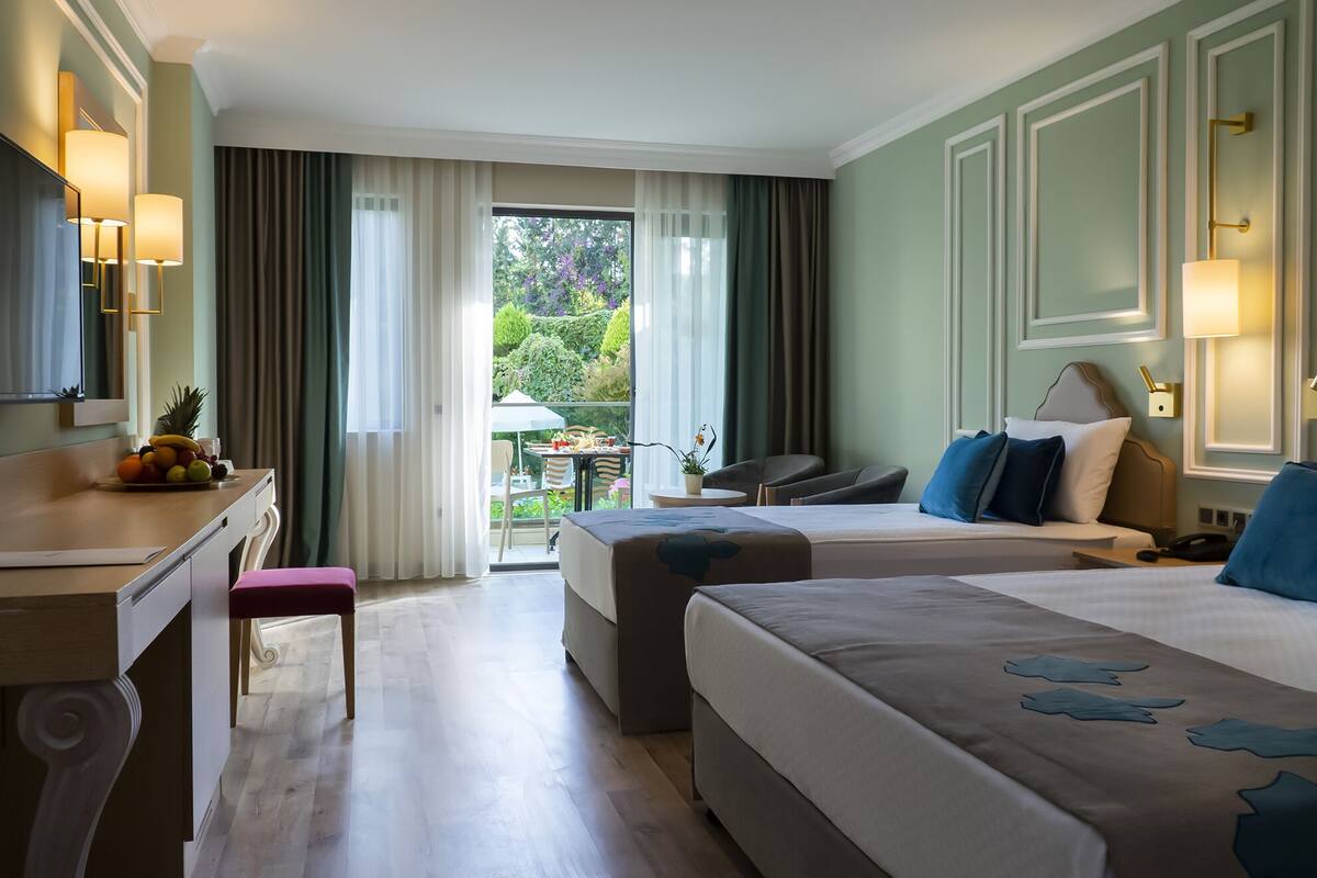 A hotel room with two beds, a desk, a chair, and a balcony with a view of greenery.