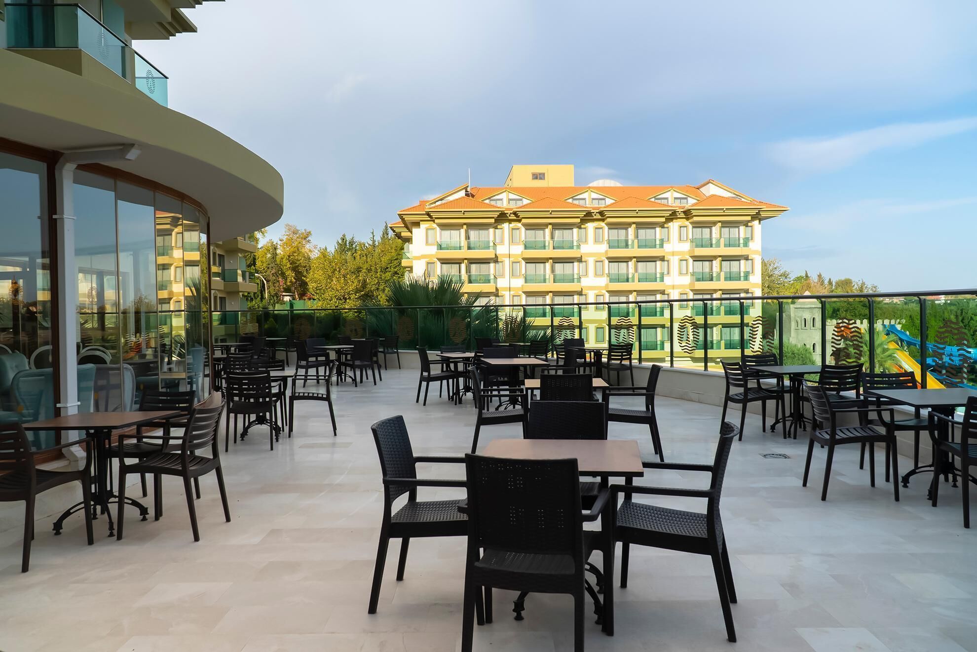 An outdoor patio with tables and chairs, overlooking a building with multiple balconies and a colorful slide.