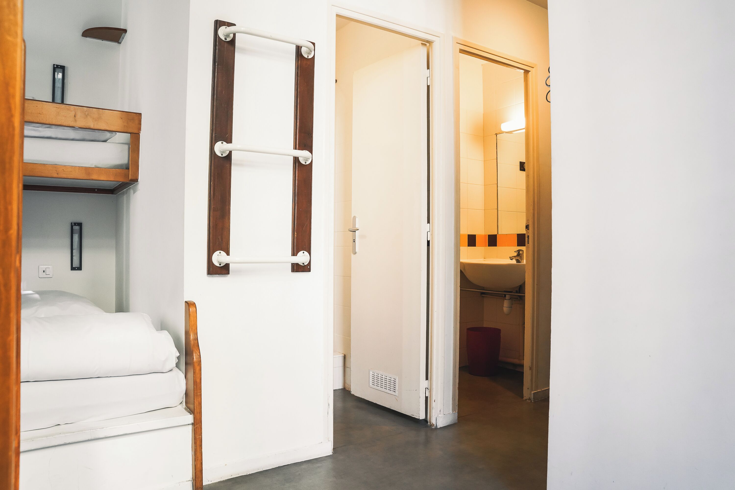 A hotel room with a bed, a wooden shelf, a towel rack, and a bathroom visible through an open door.