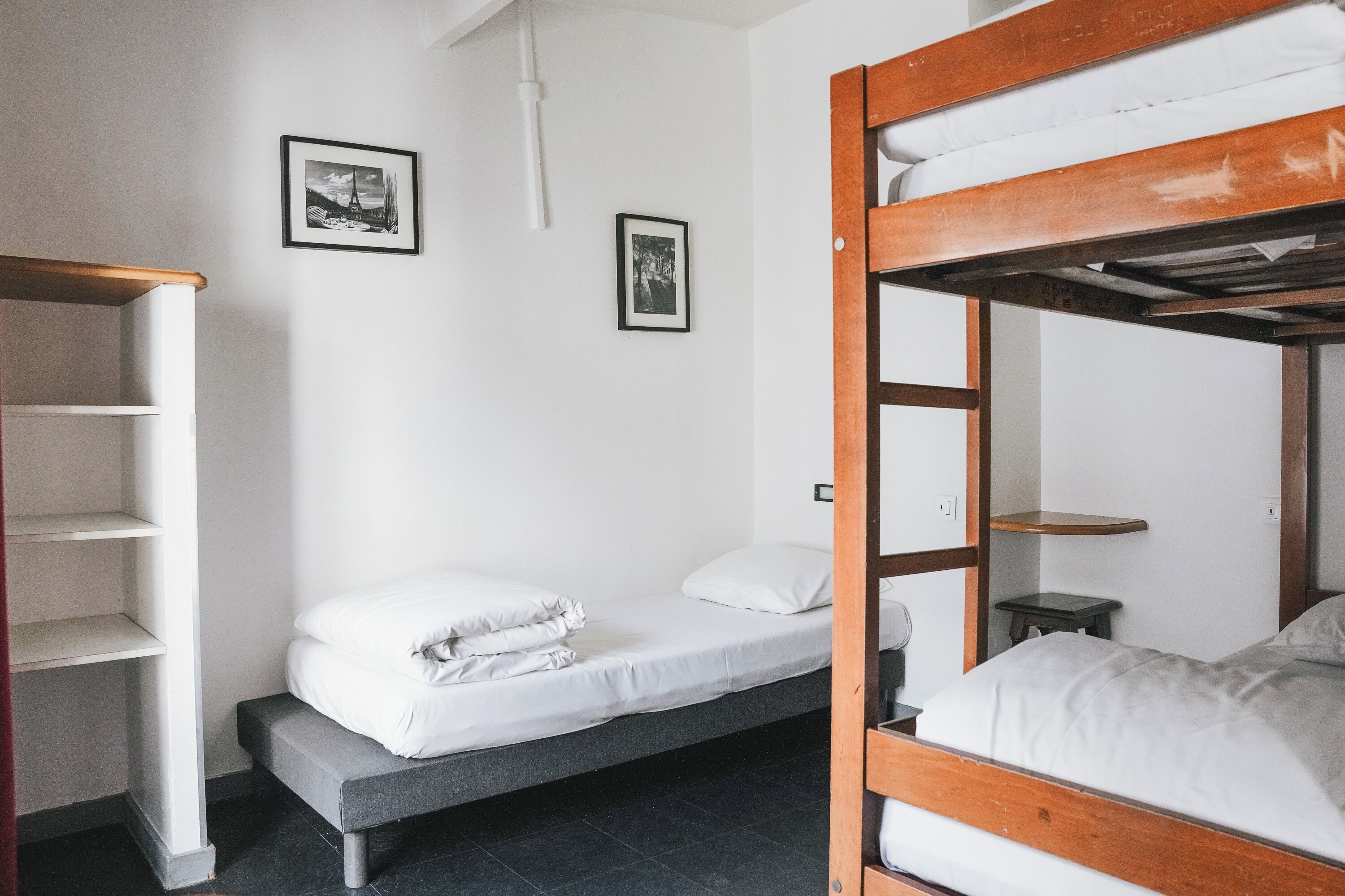 A dormitory room with two single beds, a wooden bunk bed, a small table, and a shelf unit.