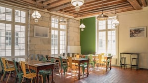 A dining area with hexagonal patterned flooring, wooden tables and chairs, and large windows with a view of buildings outside. - MIJE Marais - Hostel (Paris)