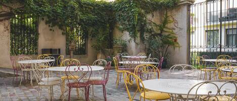 An outdoor patio with tables and chairs, surrounded by a stone floor and a wall with climbing plants.