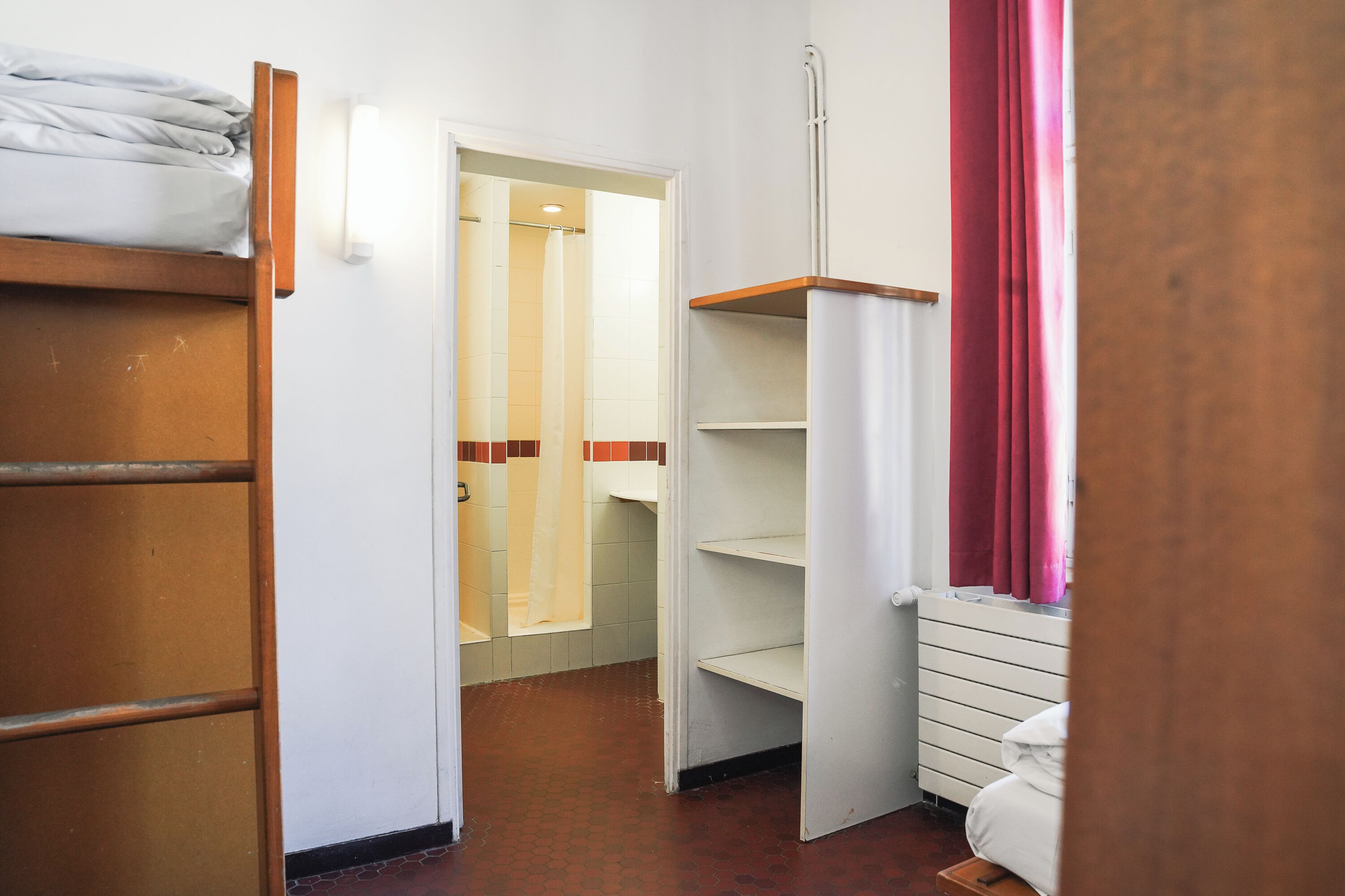 Een hotelkamer met een houten stapelbed, een witte kast, een witte radiator en een raam met rode gordijnen.