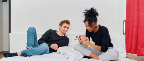 Dos personas sentadas en una cama en una habitación con pared blanca y cortina roja.
