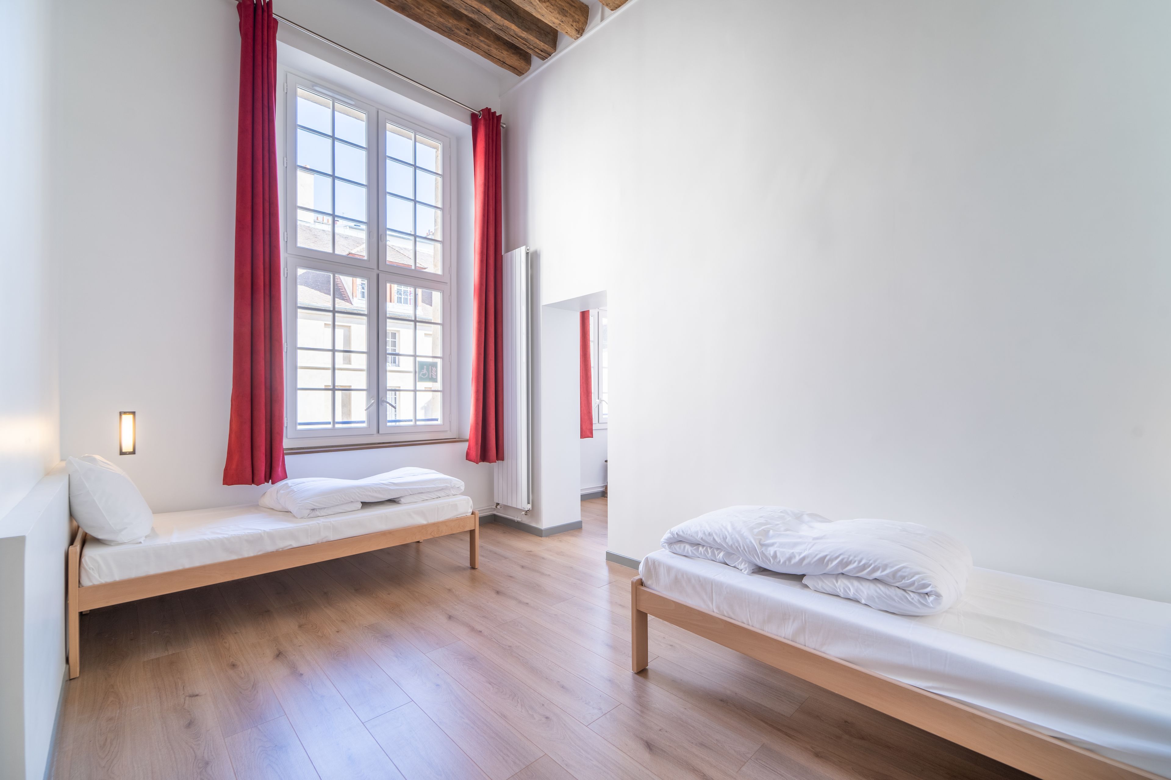 Ein Zimmer mit zwei Holzbetten, einem Fenster mit roten Vorhängen und einer weißen Wand mit Lichtschalter.