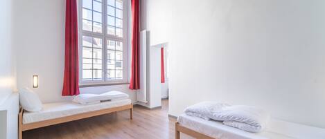 Ein Zimmer mit zwei Holzbetten, einem Fenster mit roten Vorhängen und einer weißen Wand mit Lichtschalter.