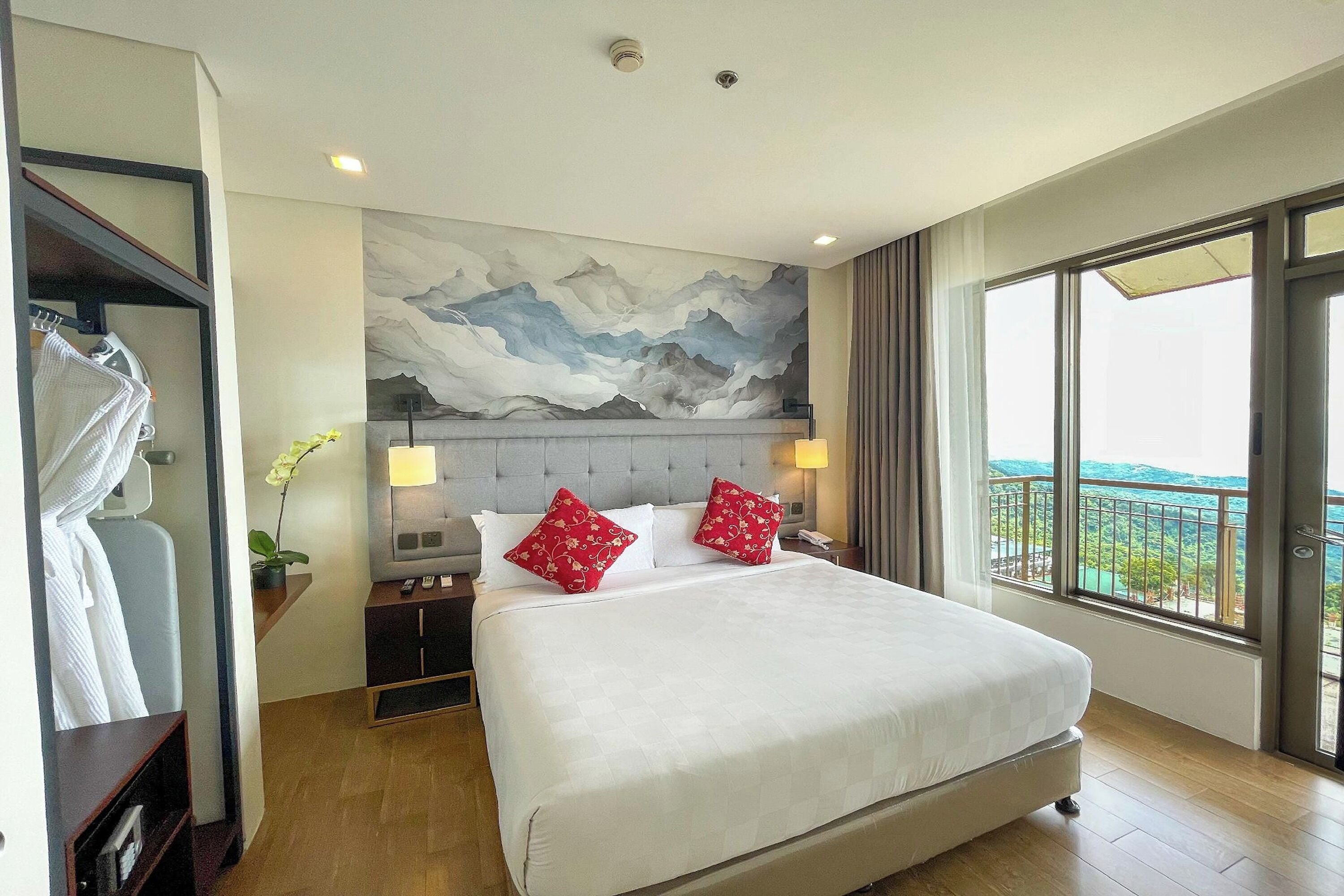 A hotel room with a large bed, a view of the outdoors, a painting on the wall, and a nightstand with a lamp.