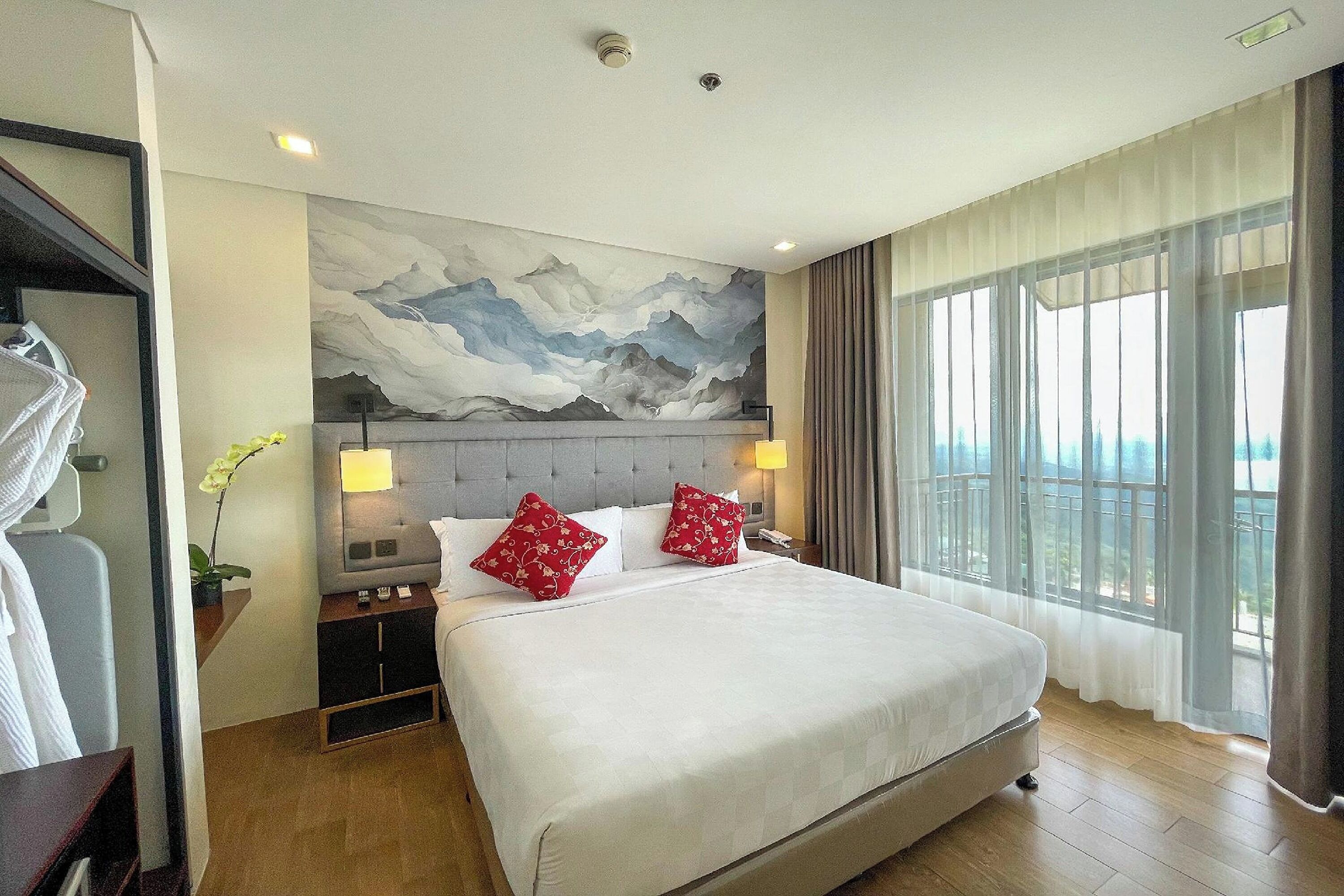 A hotel room with a large bed, a headboard with a mountain painting, two bedside lamps, a nightstand, a wardrobe, and a large window with curtains.