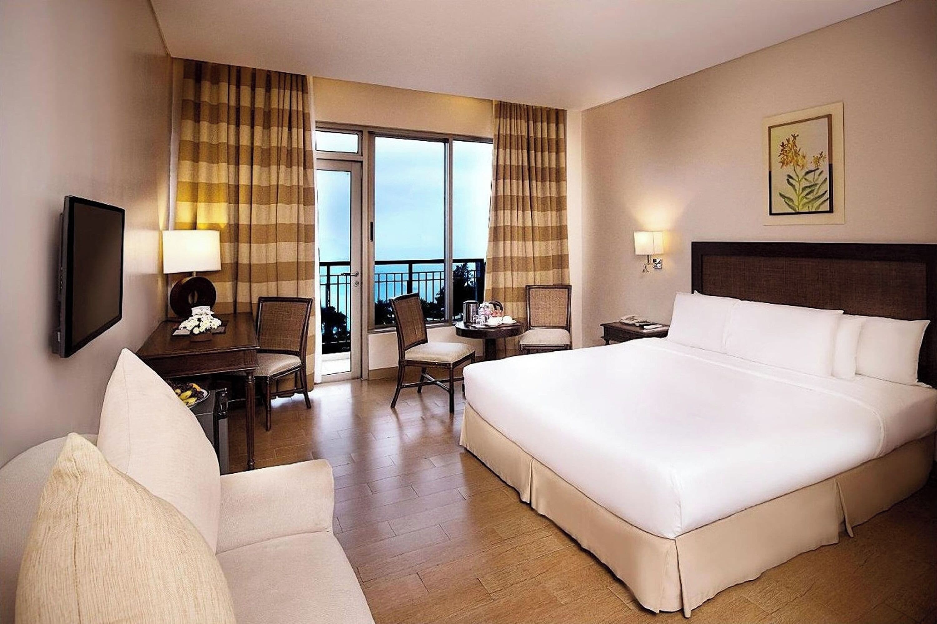 A hotel room with a large bed, a desk with a chair, a television, and a balcony with curtains.