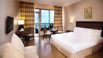 A hotel room with a large bed, a desk with a chair, a television, and a balcony with curtains.