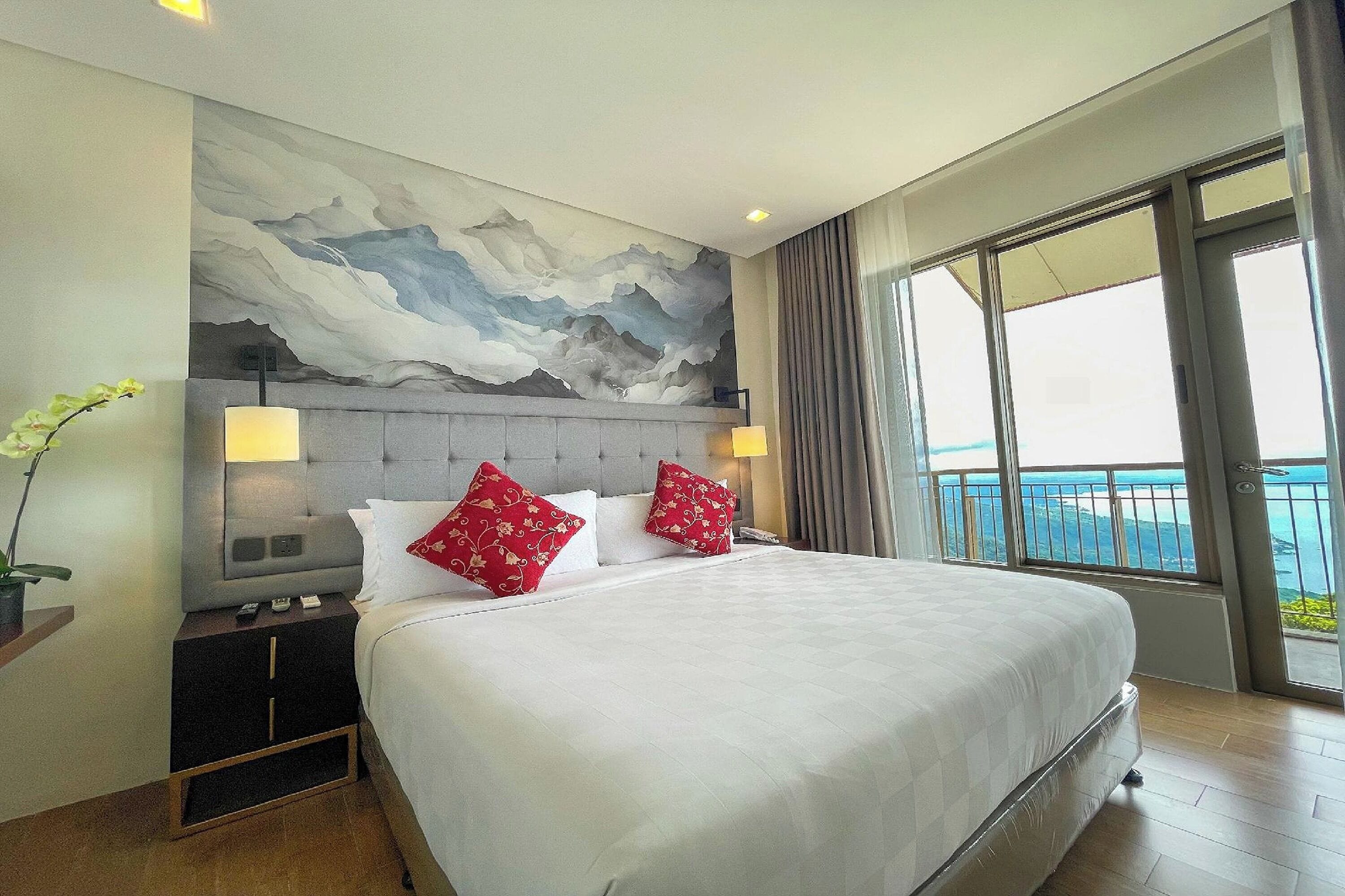 A hotel room with a large bed, two red pillows, a nightstand, a lamp, and a view of the sea through large windows.