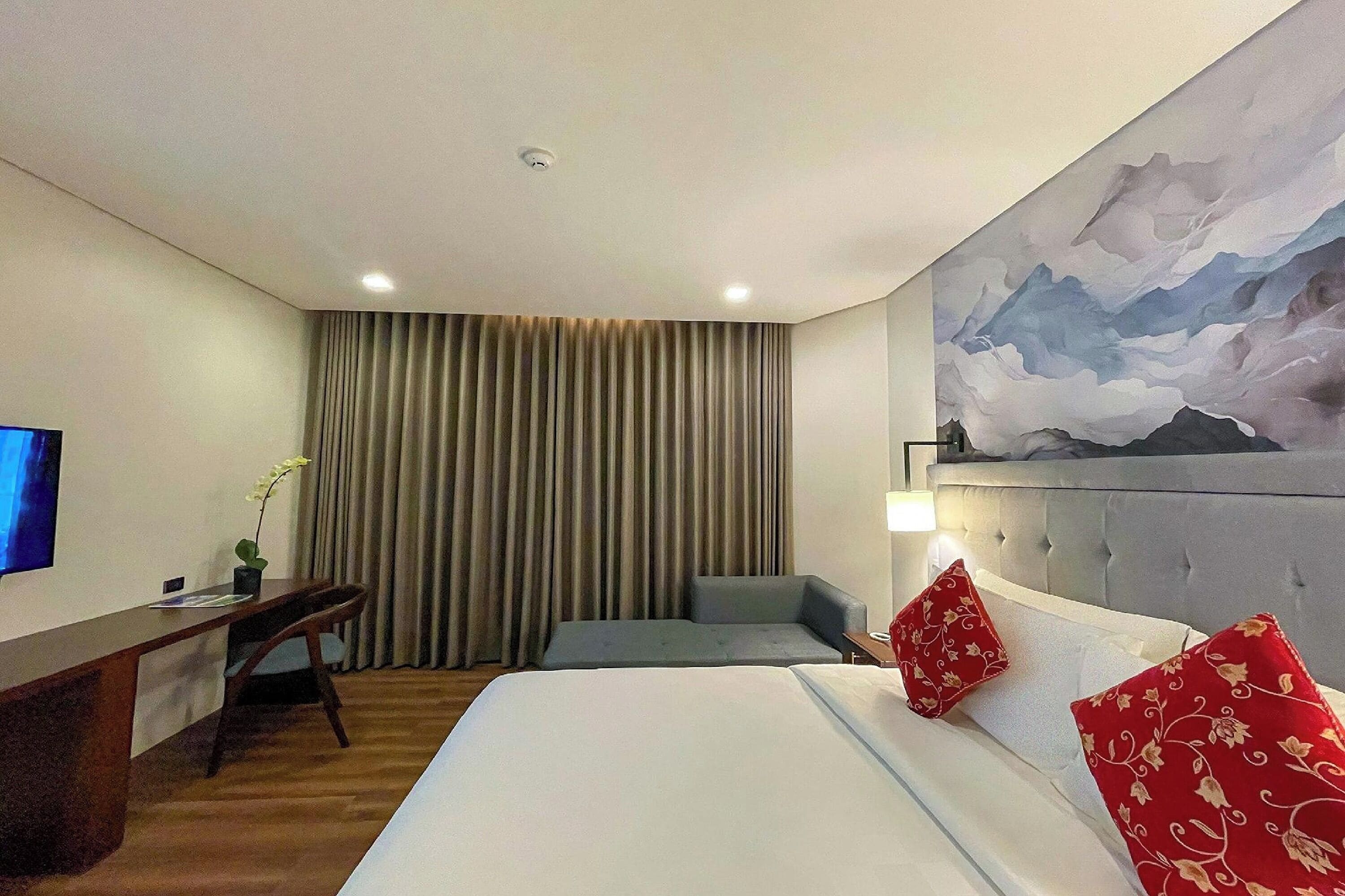 A hotel room with a bed, a desk, a chair, a TV, and a painting on the wall.