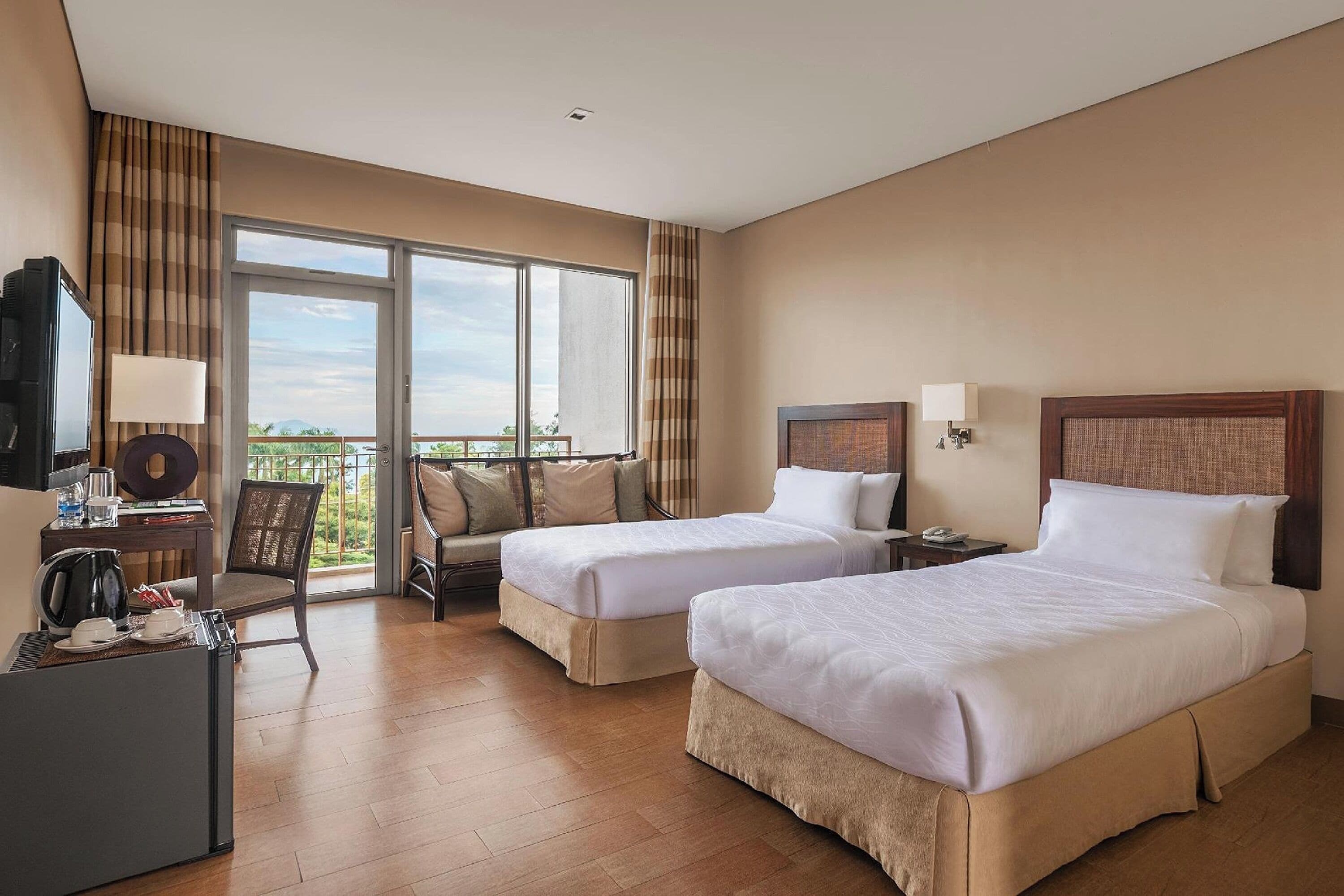 A hotel room with two beds, a desk with a television, a chair, and a balcony with a view.