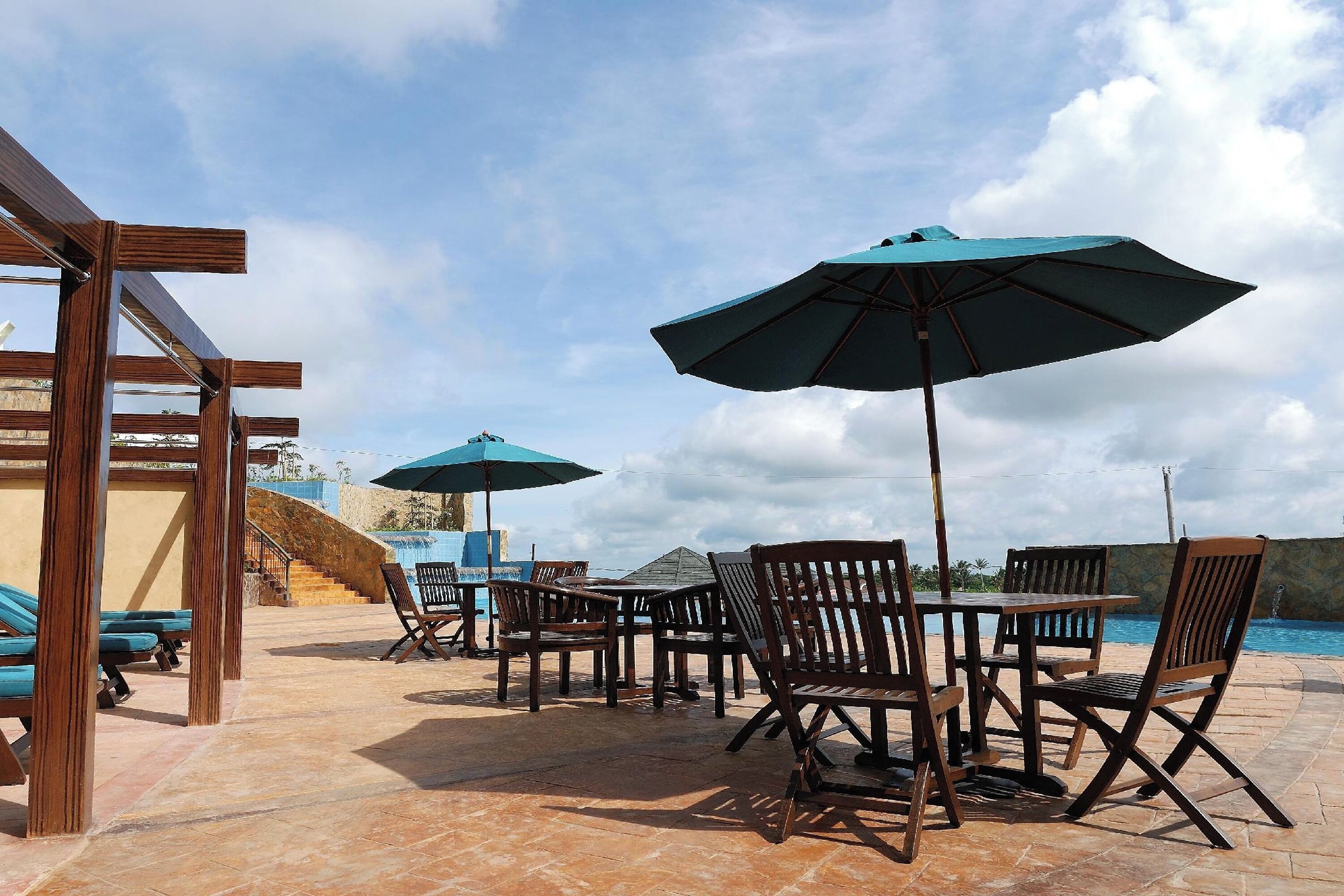 An outdoor seating area with wooden tables and chairs, blue umbrellas, and a pool in the background.