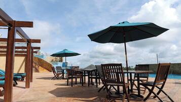 An outdoor seating area with wooden tables and chairs, blue umbrellas, and a pool in the background.